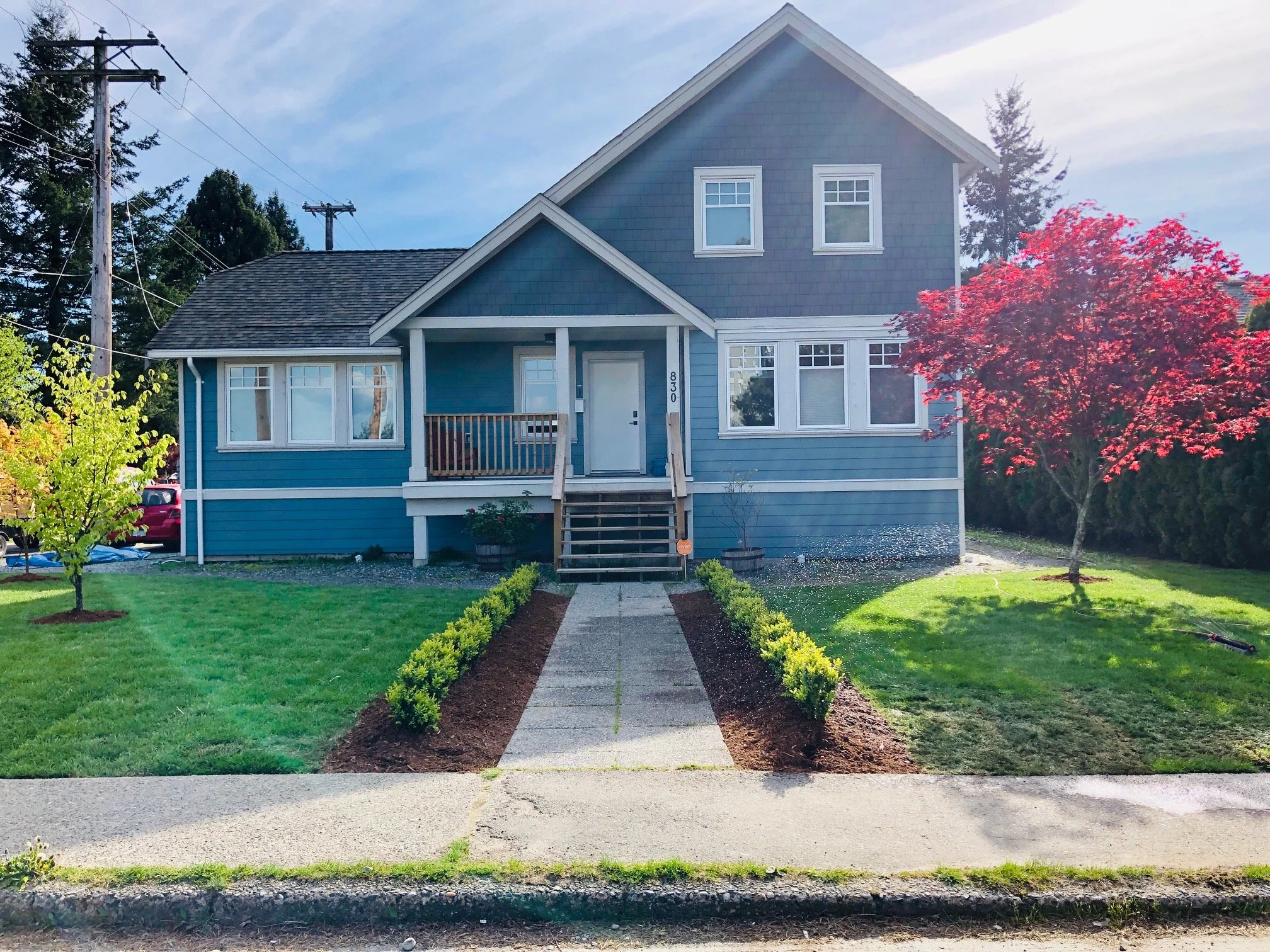 Front view of a two-story blue house with a porch, steps leading to the entrance, and a well-maintained lawn with two trees and landscaped bushes.