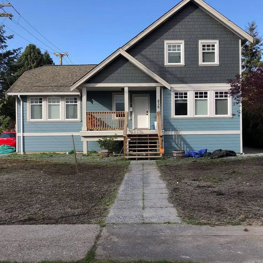 Front view of a blue, two-story house with a small porch, wooden stairs, and white trim. There are five windows visible on the house, and the house number 830 is near the front door. The yard is mostly bare dirt with some construction materials and a
