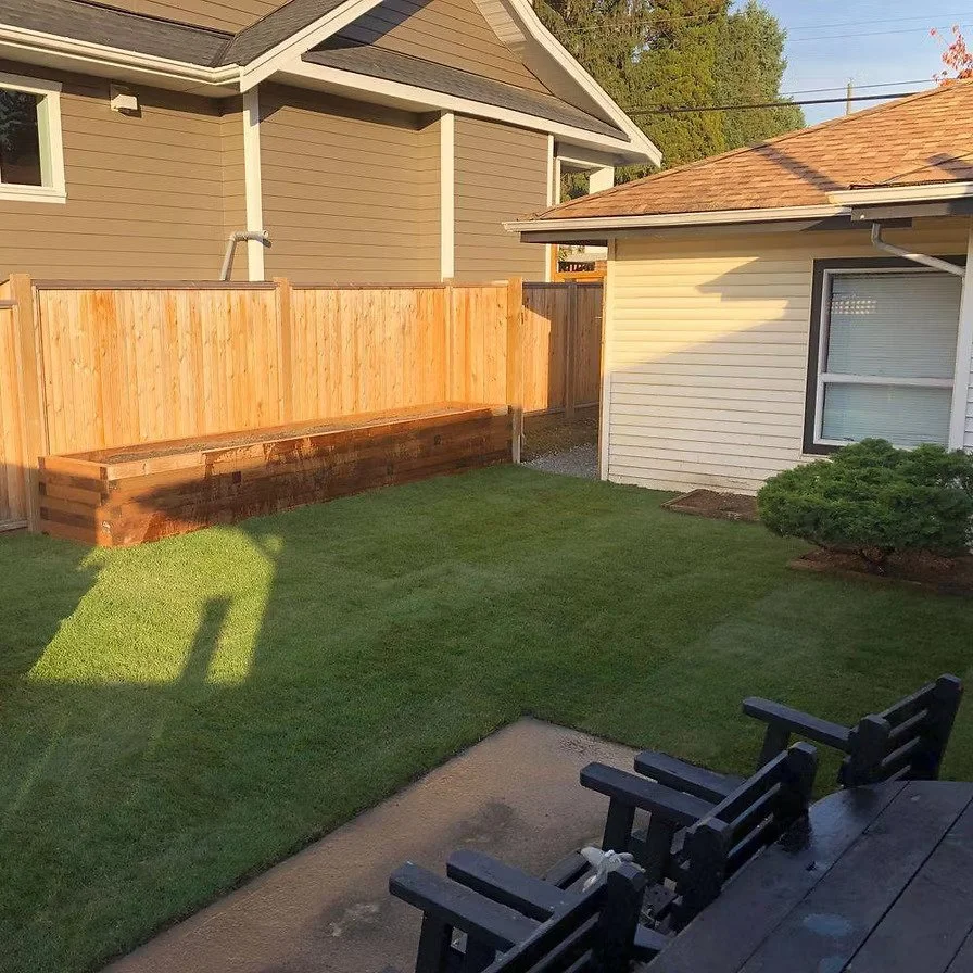 View of a backyard with a freshly built wooden fence and a planter box, green lawn, a small bush, and part of a deck with black chairs.