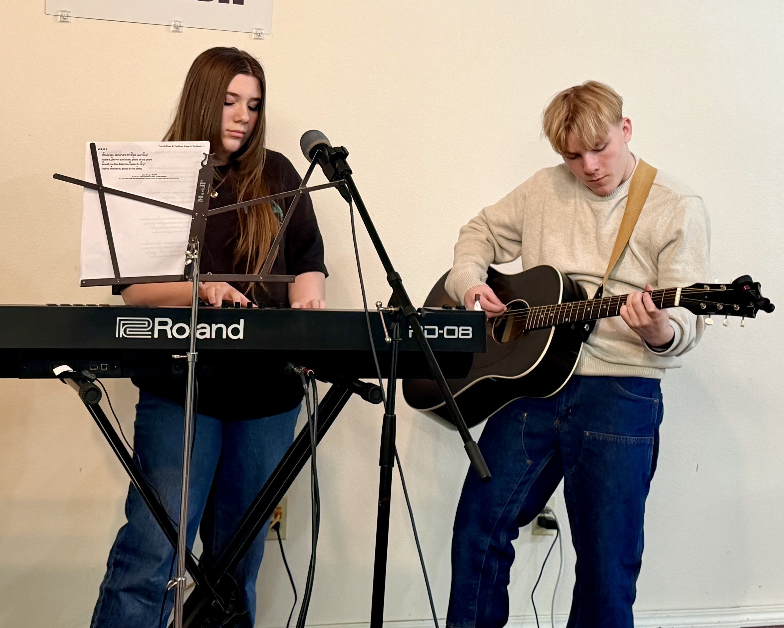 A young woman playing a keyboard and a young man playing an acoustic guitar performing together in a room.