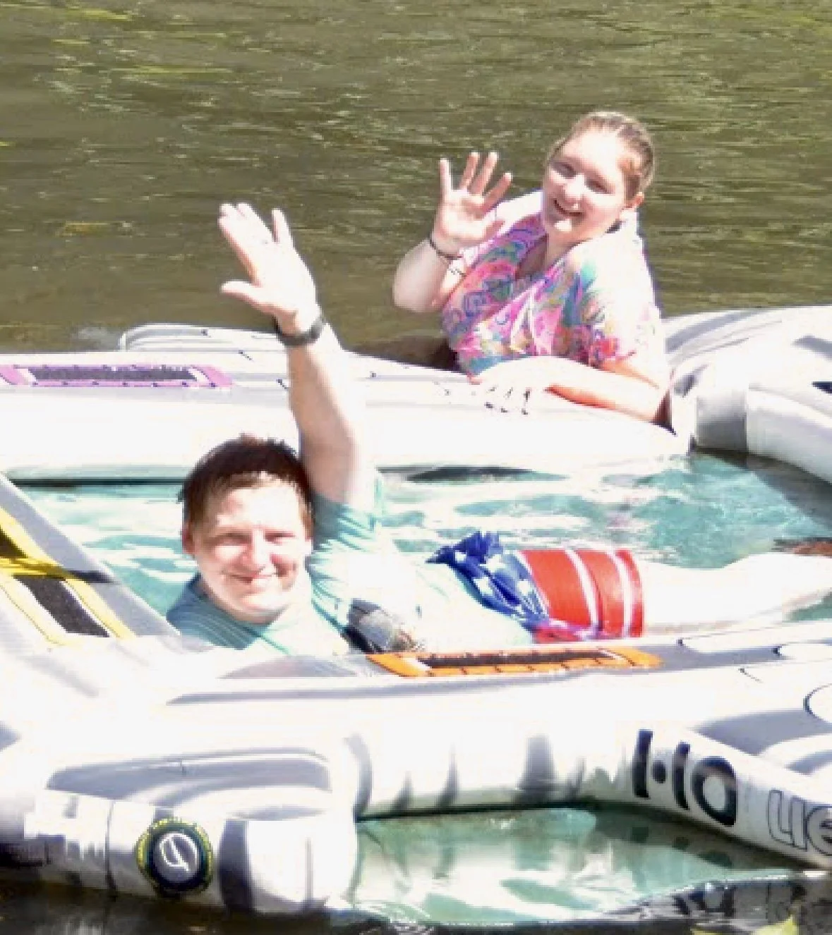 Two people in boats on water, waving and smiling.