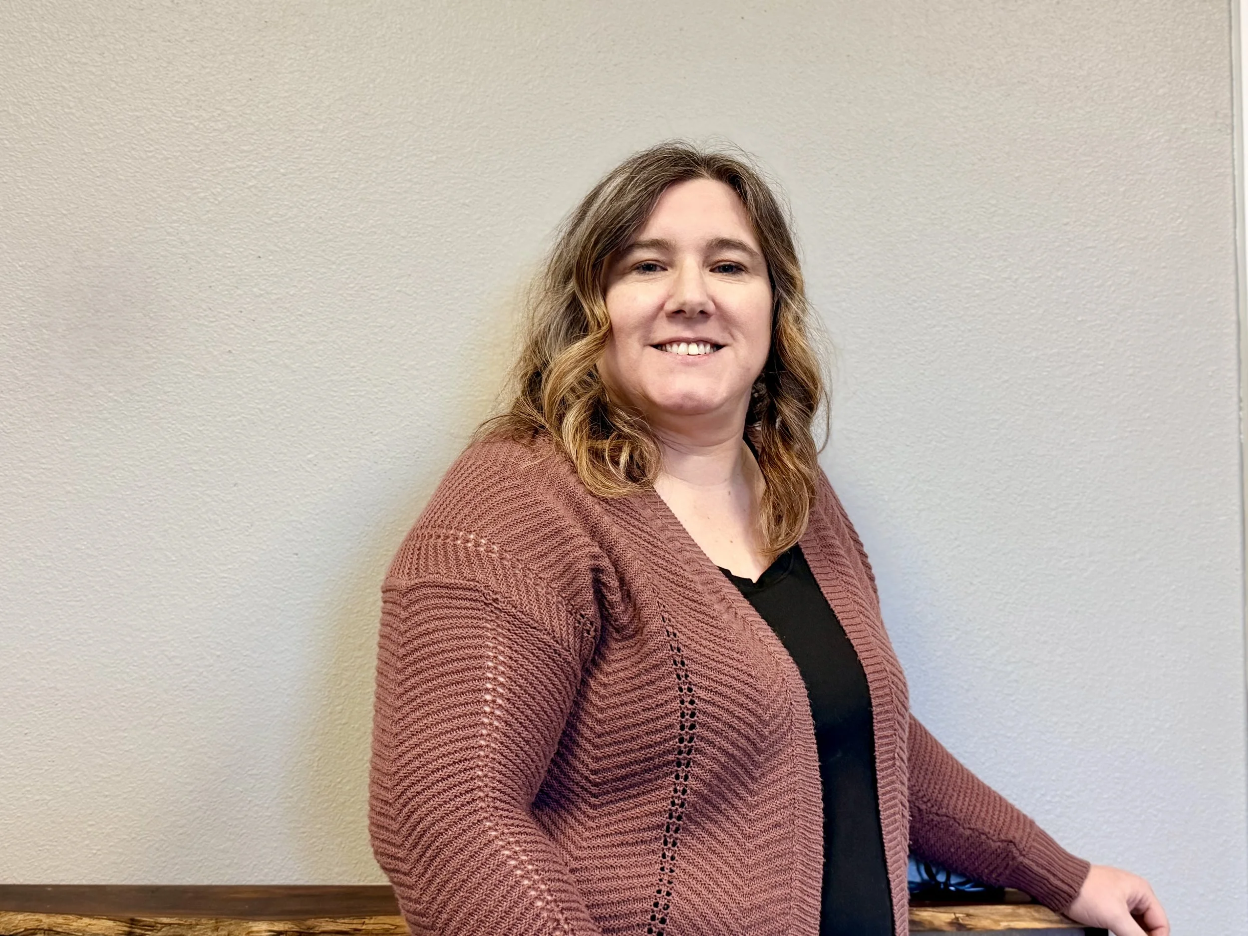 A woman with shoulder-length wavy hair smiling, wearing a mauve cardigan over a black top, standing against a light-colored textured wall.
