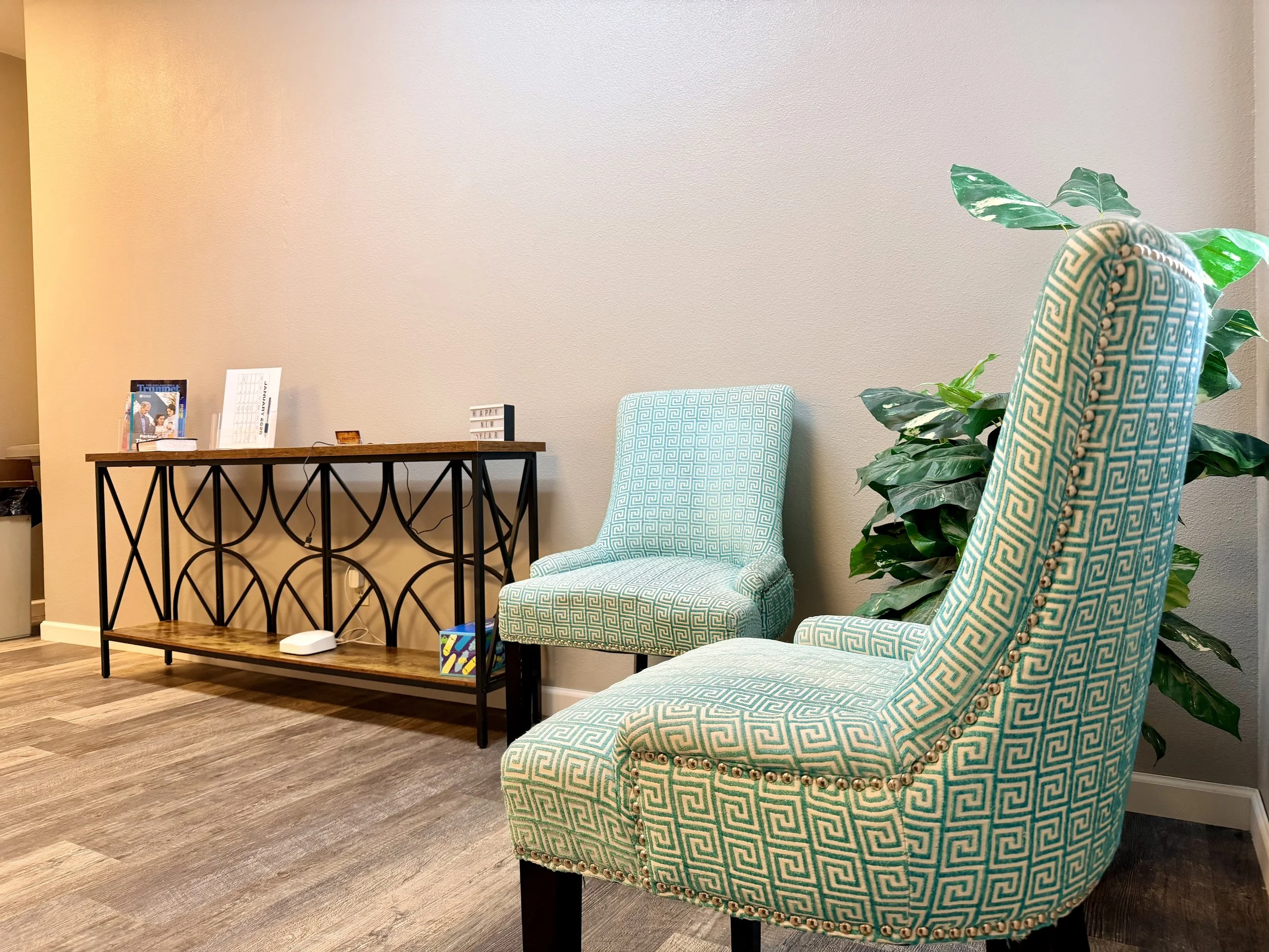 A waiting area with two patterned chairs, a wooden console table, and a houseplant against a beige wall.