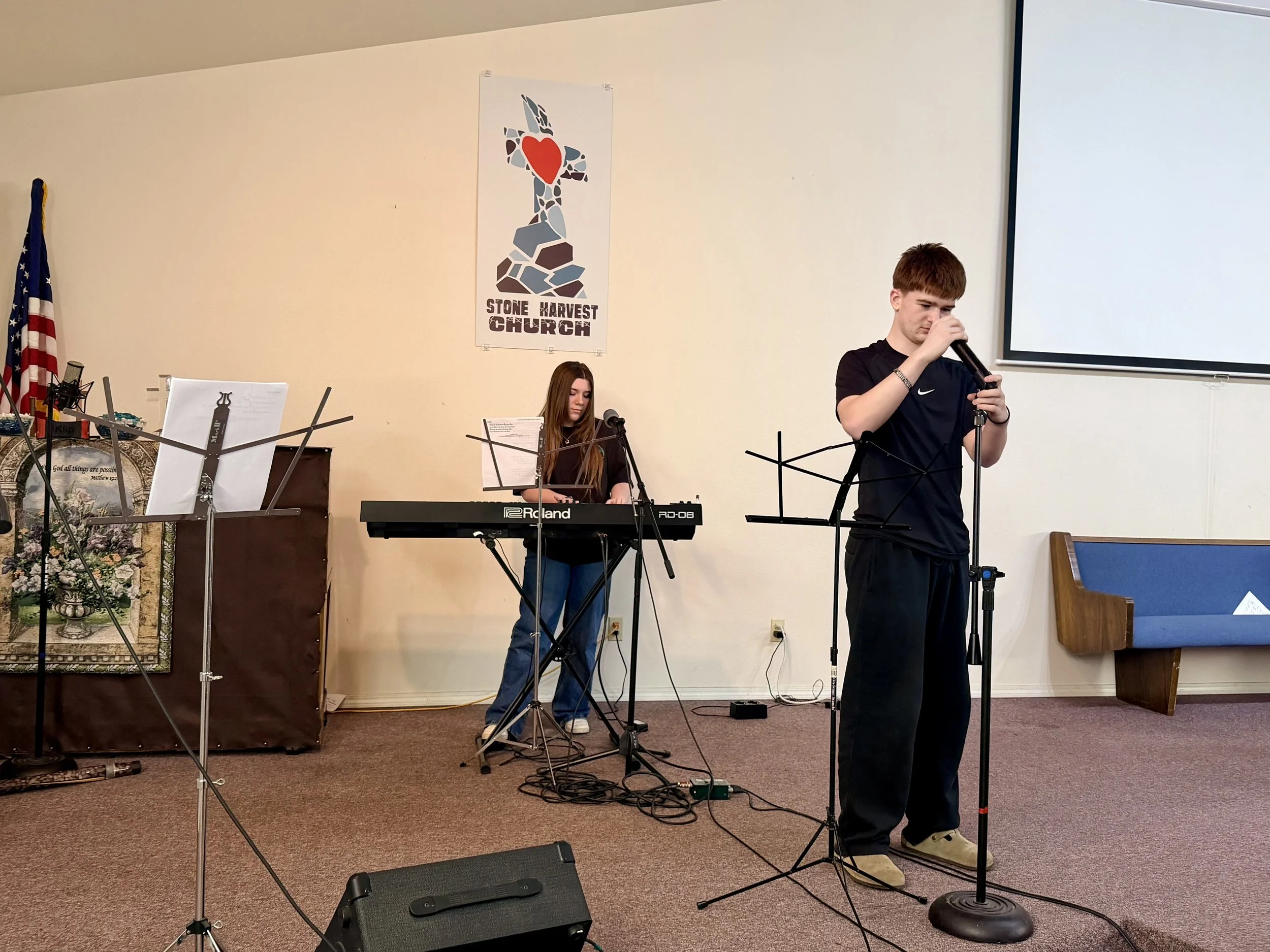 A young man singing into a microphone and a young woman playing a keyboard in a church setting with a Stone Harvest Church banner and American flag in the background.