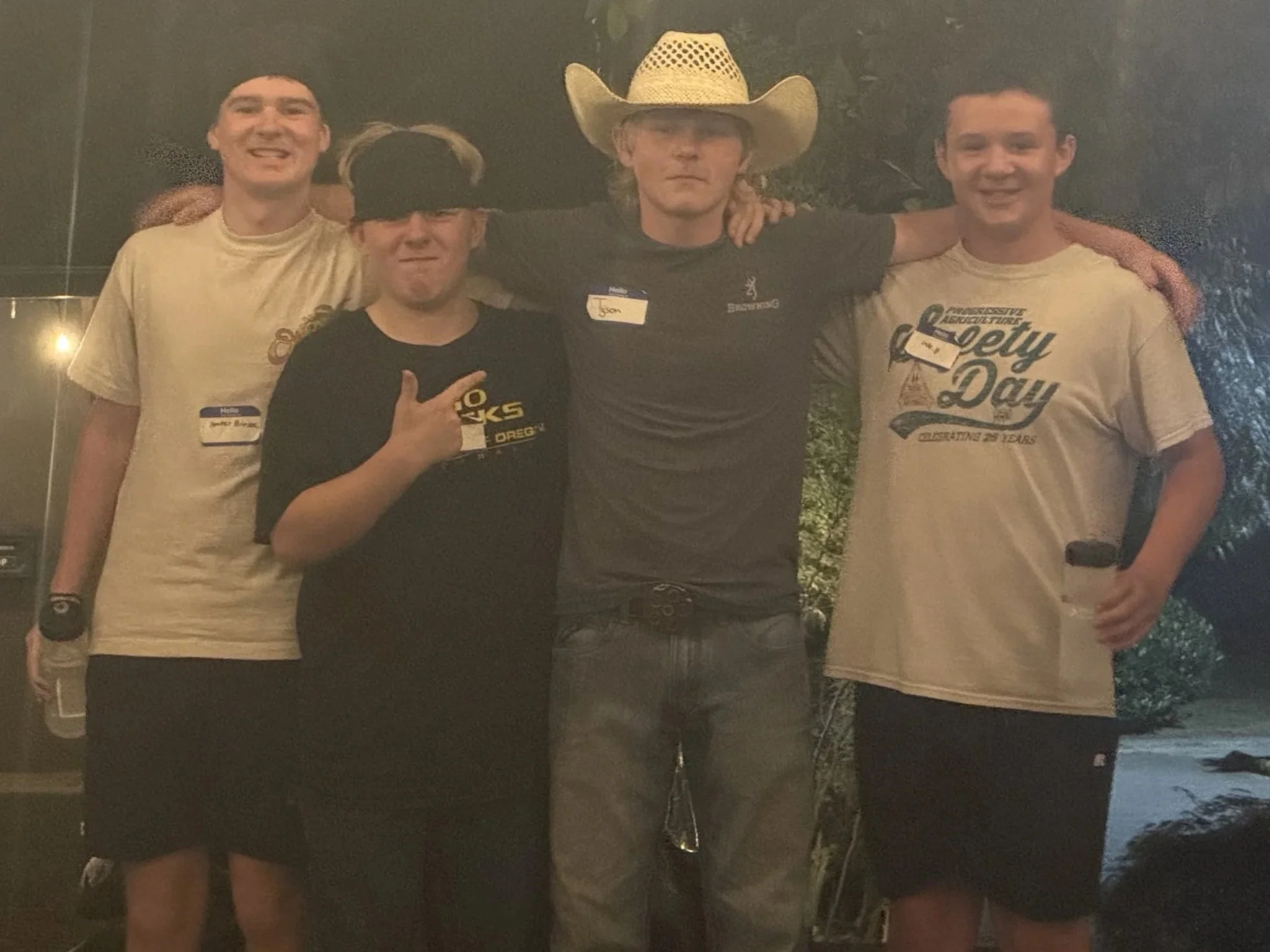 A group of four young men standing outdoors at night, with some trees visible in the background. They are smiling and posing with their arms around each other. Two of them are holding water bottles. One man is wearing a cowboy hat and a black T-shirt
