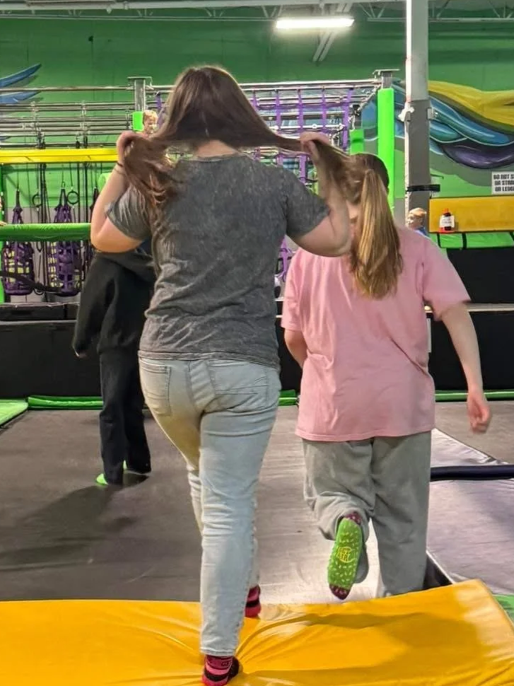 Two girls with long hair at an indoor trampoline park. The girl on the left has dark hair and is wearing a gray t-shirt and light jeans. The girl on the right has light brown hair and is wearing a pink shirt and gray sweatpants. They're walking on a 
