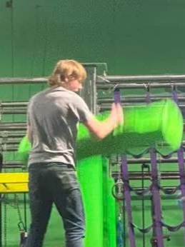 A person with blonde hair wearing a gray shirt and jeans, operating a large green pipe or tube in a green-walled industrial setting.