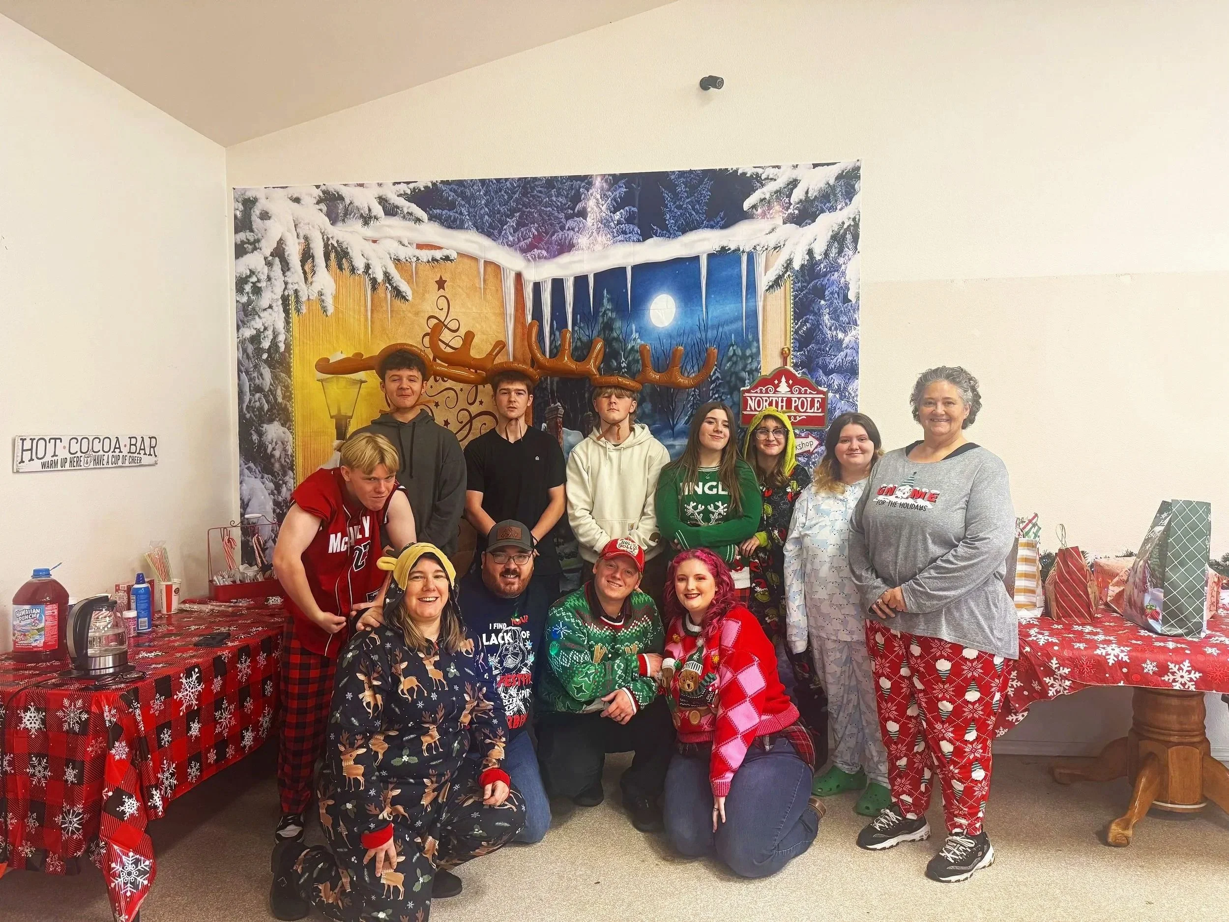 A group of people dressed in Christmas-themed pajamas and sweaters posing in front of holiday decorations, including a backdrop with a snowy night scene, reindeer antlers, and a North Pole sign, at a Christmas party.
