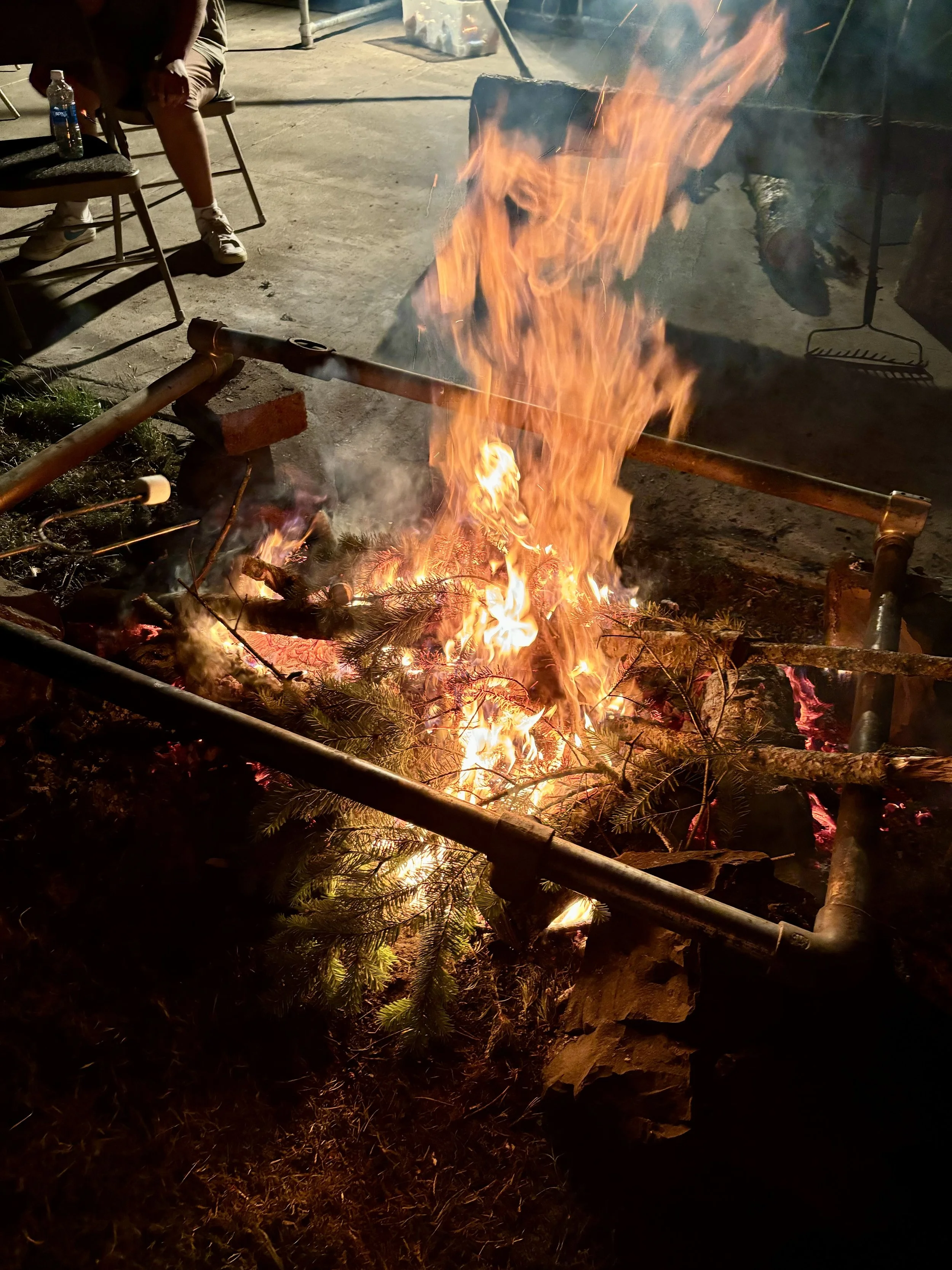 Fire burning with wood, branches, and pine needles, surrounded by metal pipes and chairs, at night.