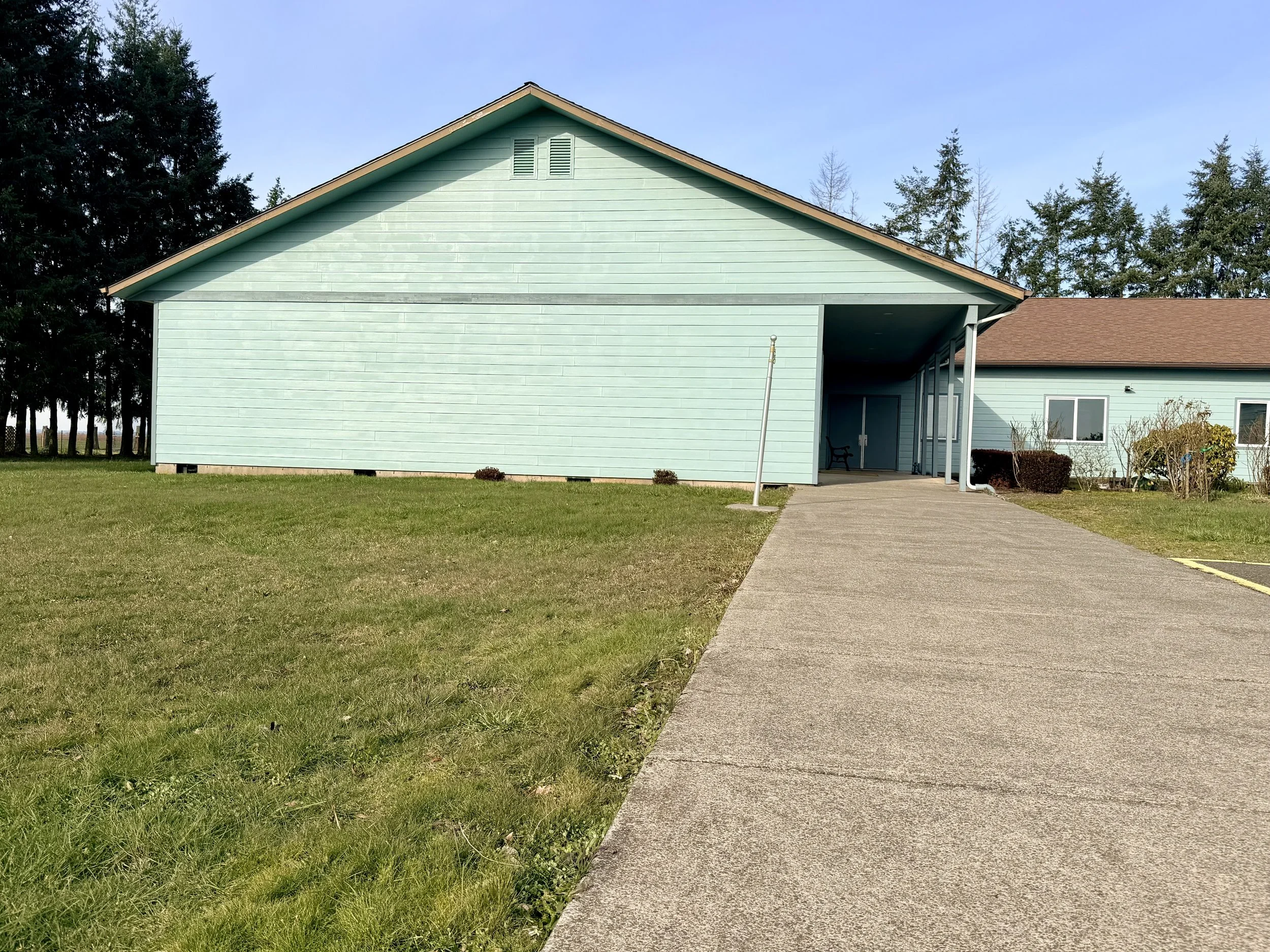 A light blue building with a brown roof and a concrete driveway leading to an entrance porch, surrounded by grass and trees.