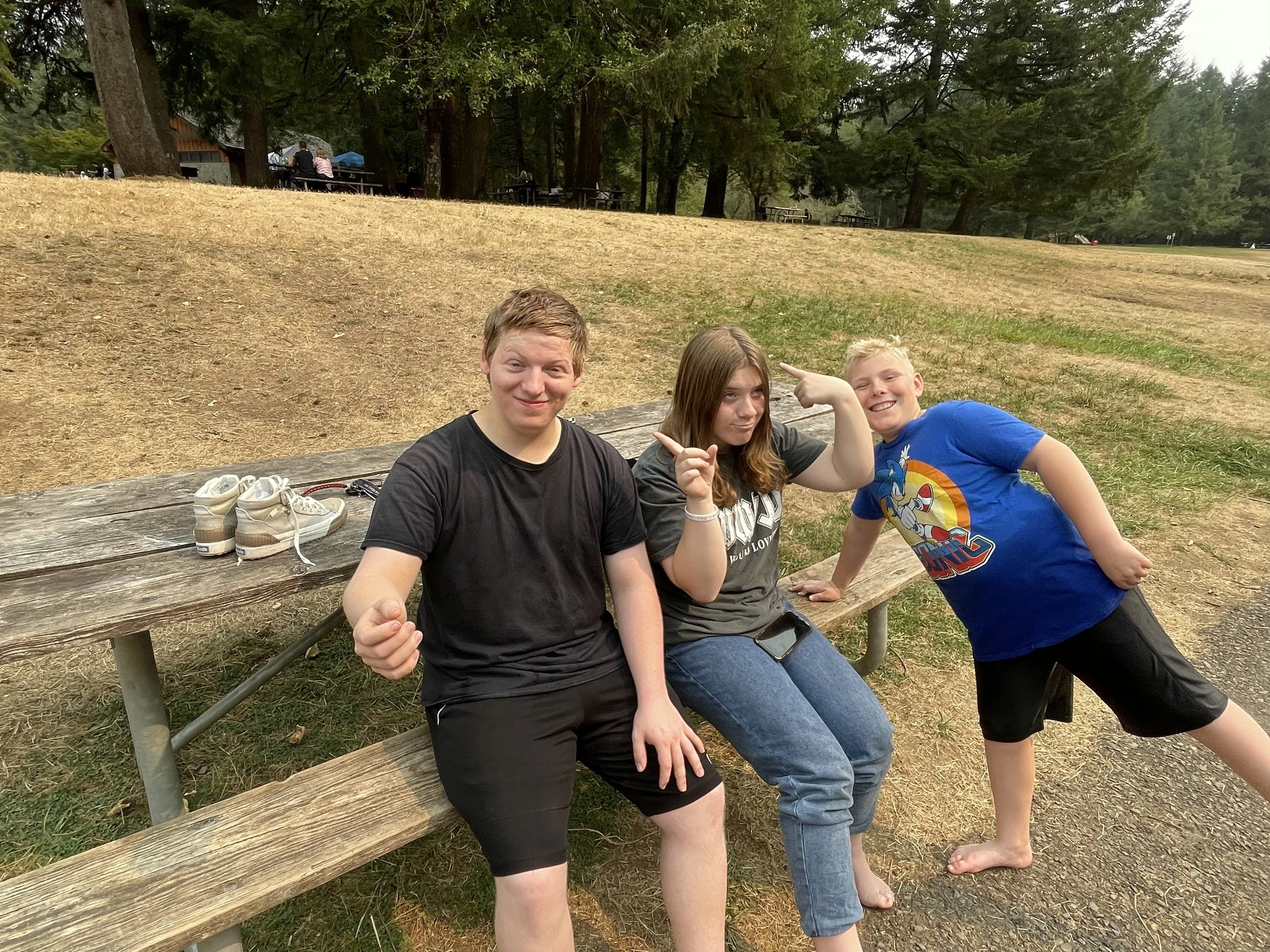 Three children sitting and standing on a wooden bench outdoors, smiling and posing playfully, with shoes on the bench beside them, in a park with trees and grass in the background.