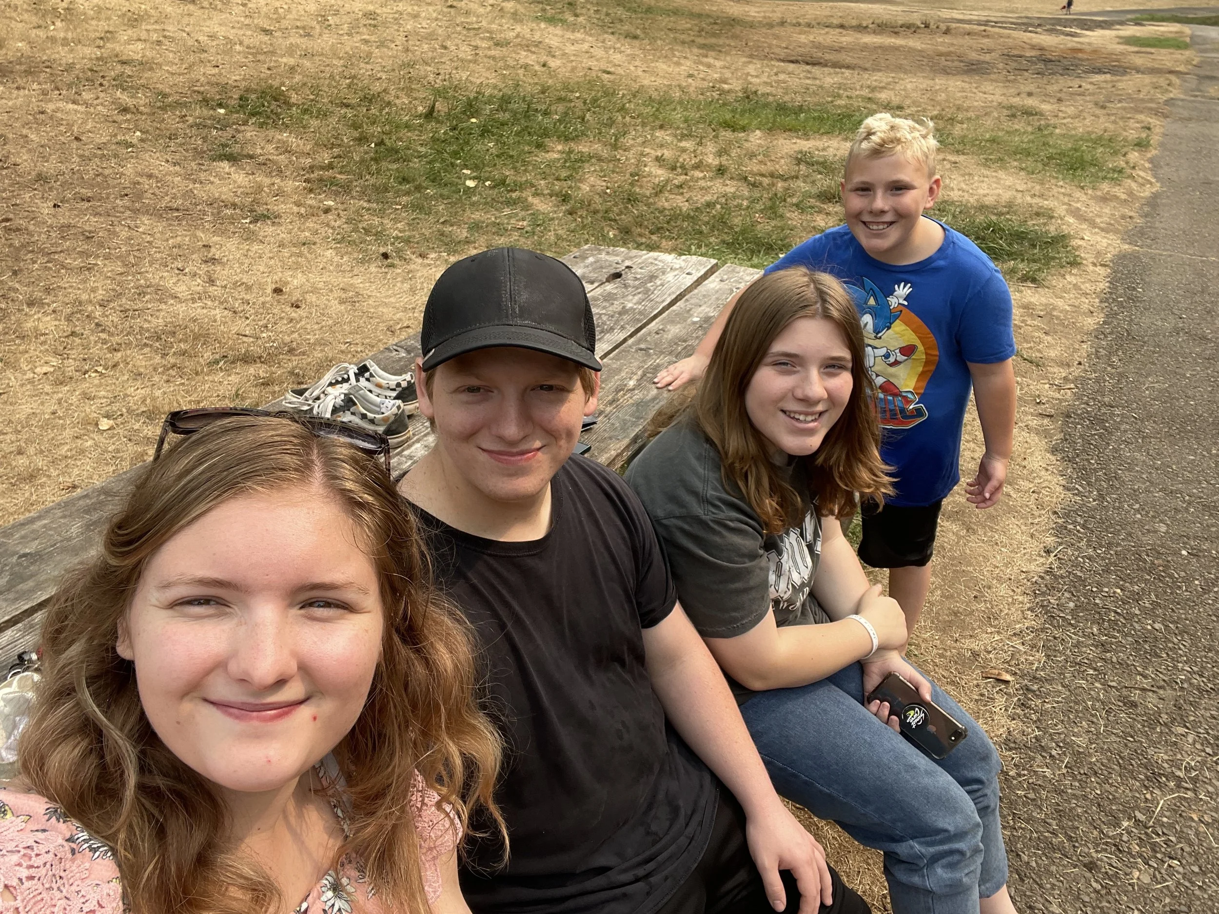 Four young friends sitting on a park bench outdoors, smiling at the camera. One girl is in the front with sunglasses on her head, a boy with a black cap is next to her, a girl with long hair is to his right, and a boy wearing a blue Sonic the Hedgeho