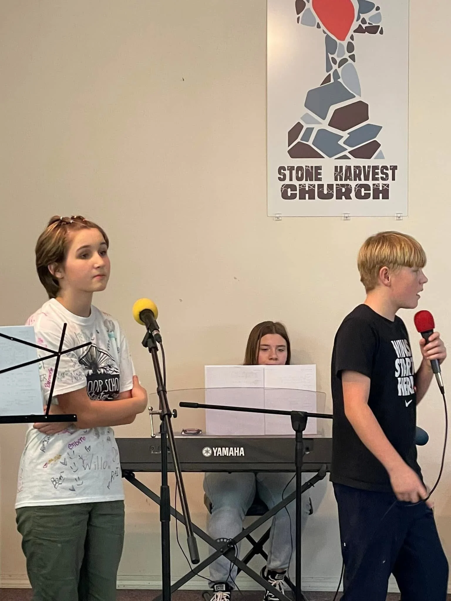 Three young children performing or practicing at Stone Harvest Church, with one girl on the left in a signed white T-shirt, a boy on the right holding a red microphone, and a girl in the middle playing a Yamaha keyboard.