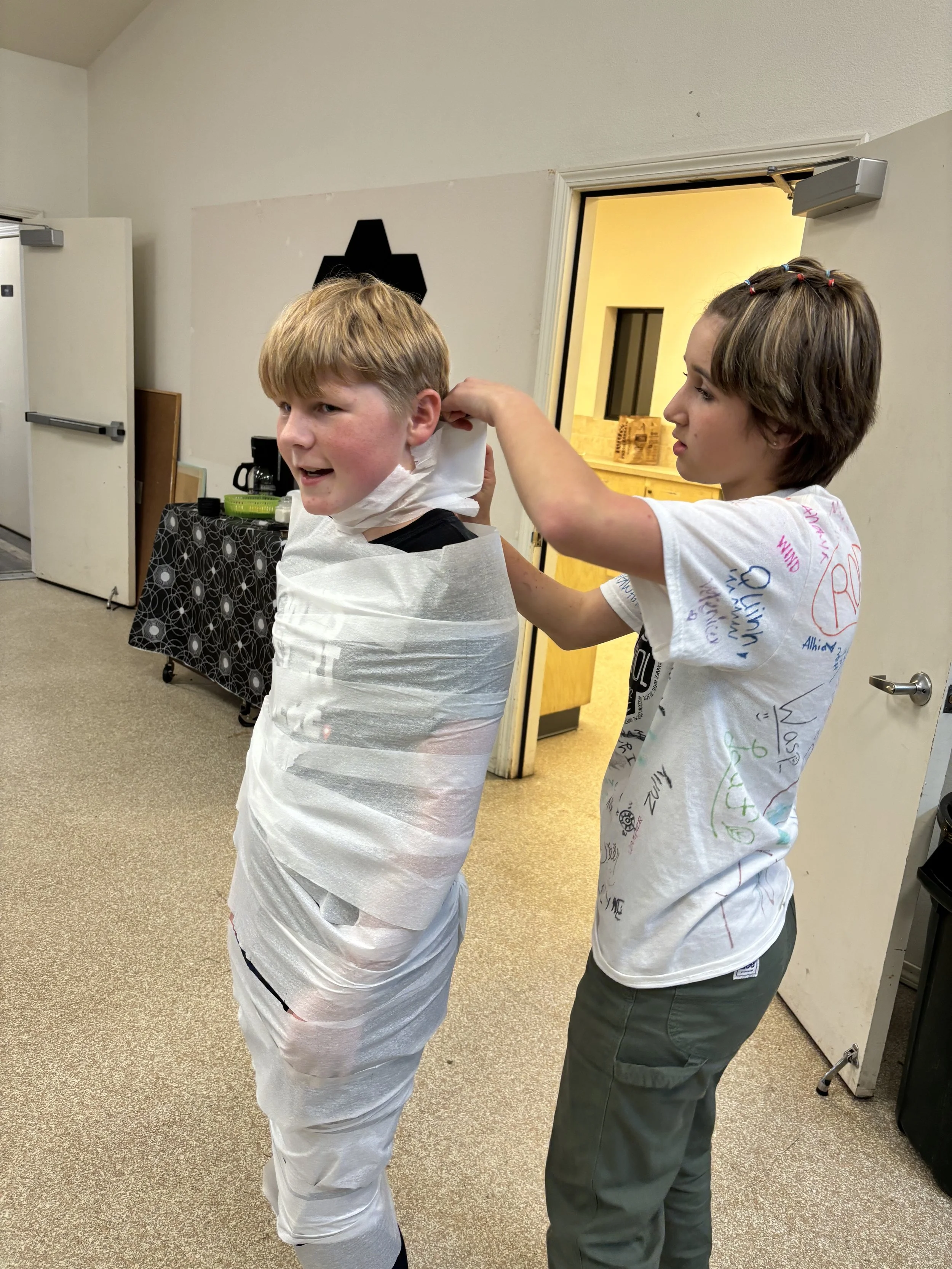 A young boy is wrapped in white toilet paper by a girl in a room with a beige carpet and light walls, as part of a fun activity.