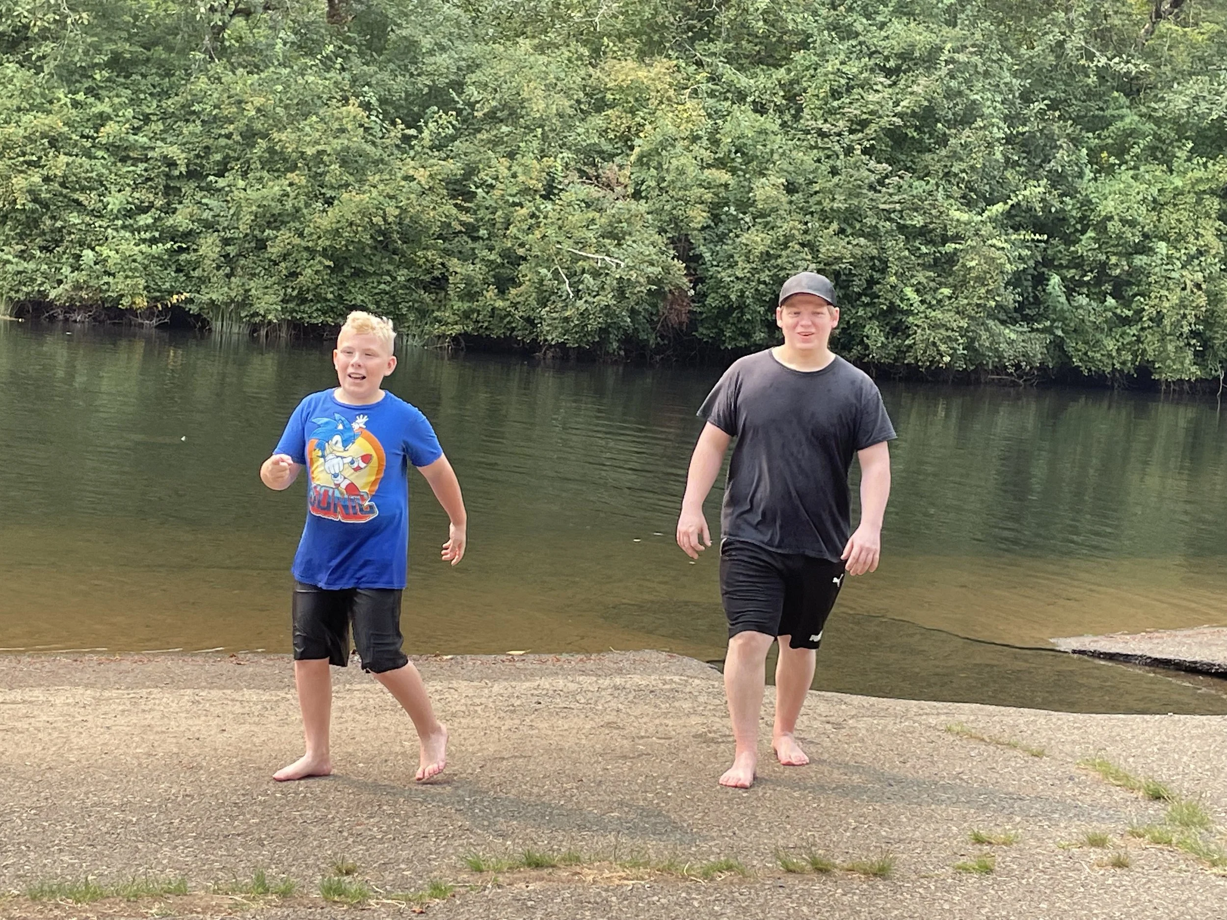 Two boys walking barefoot on a concrete path near a lake with lush green trees in the background.