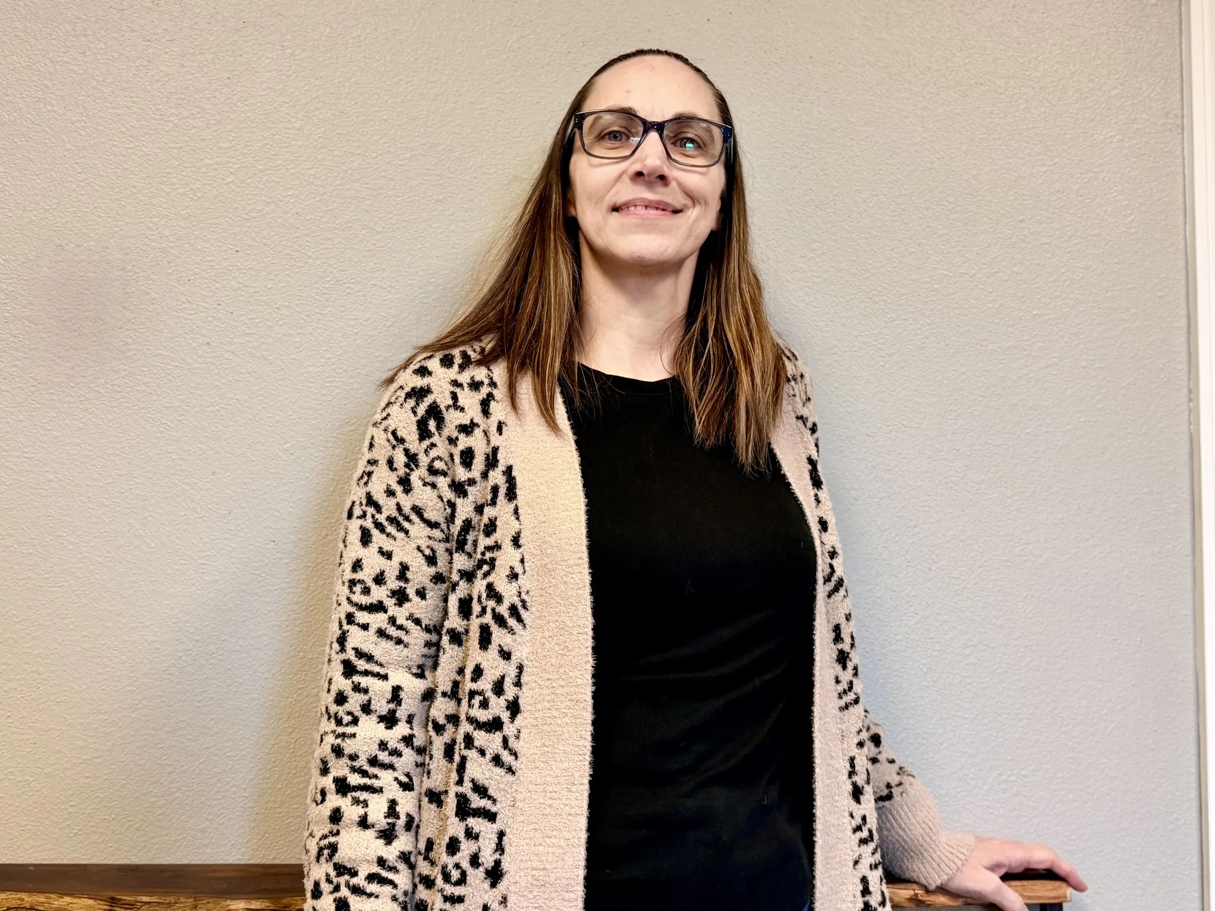 A woman with long brown hair, wearing glasses, a black shirt, and a beige leopard print cardigan, standing against a beige wall.