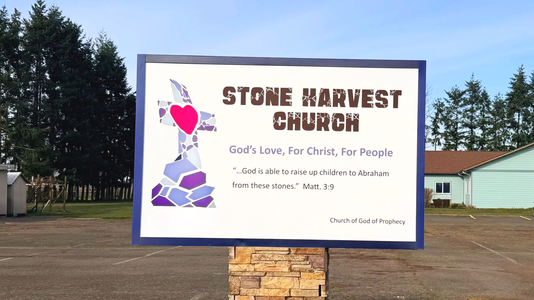 A church sign for Stone Harvest Church with a mosaic cross and a pink heart in the center. The sign includes a Bible quote from Matthew 3:9 and the church's mission statement, 'God's Love, For Christ, For People,' in a parking lot with trees and a building in the background.