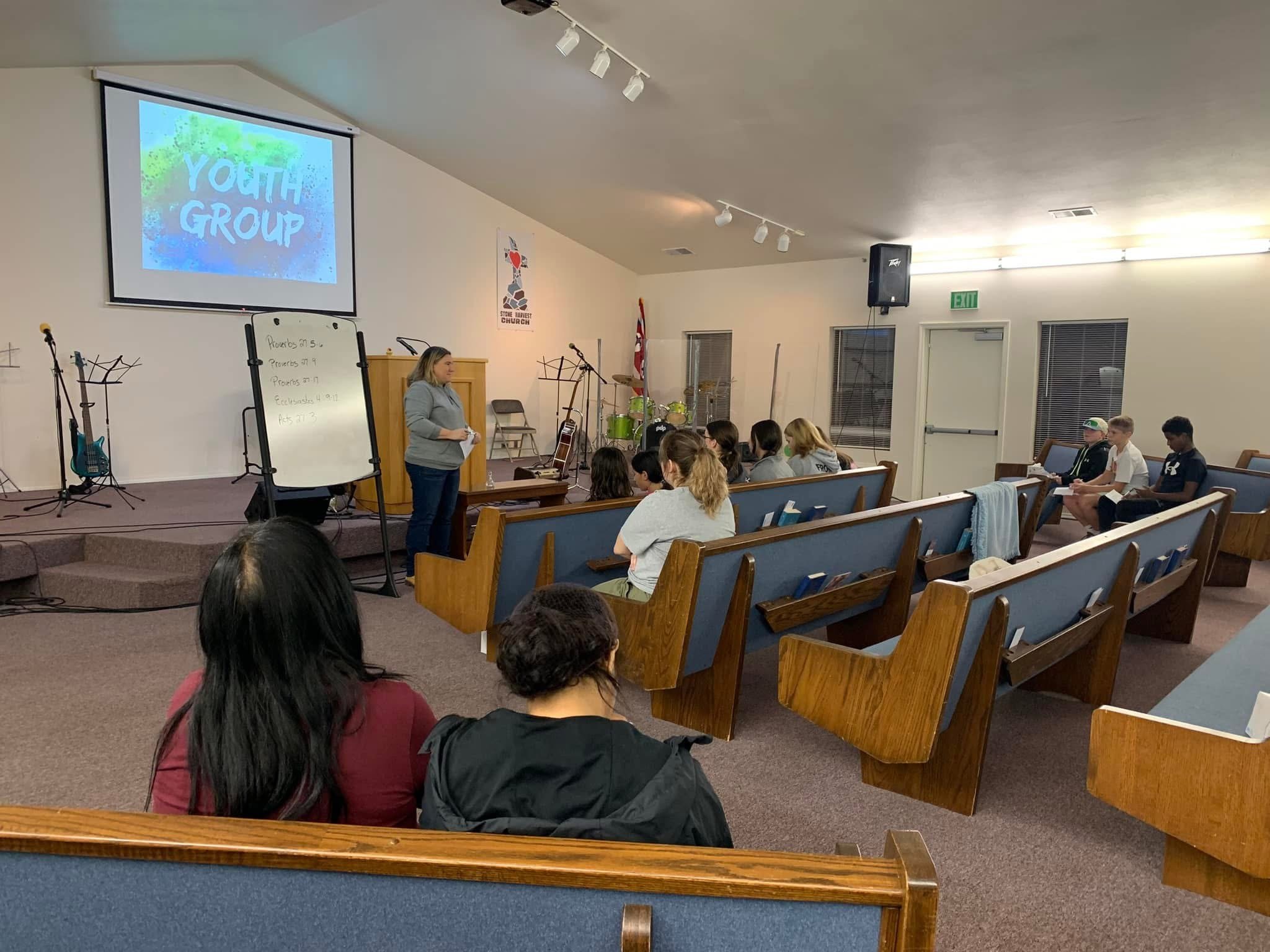 A youth group meeting in a church auditorium with a woman speaking at a podium, a whiteboard with scriptures, a large screen displaying 'YOUTH GROUP', and musical instruments around the stage.