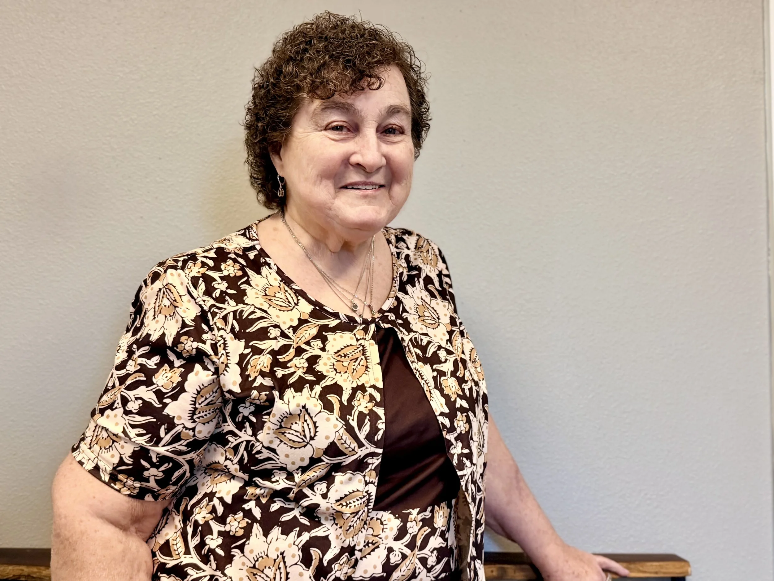 An elderly woman with short curly hair smiling, wearing a floral patterned jacket over a dark top, standing against a plain beige wall.