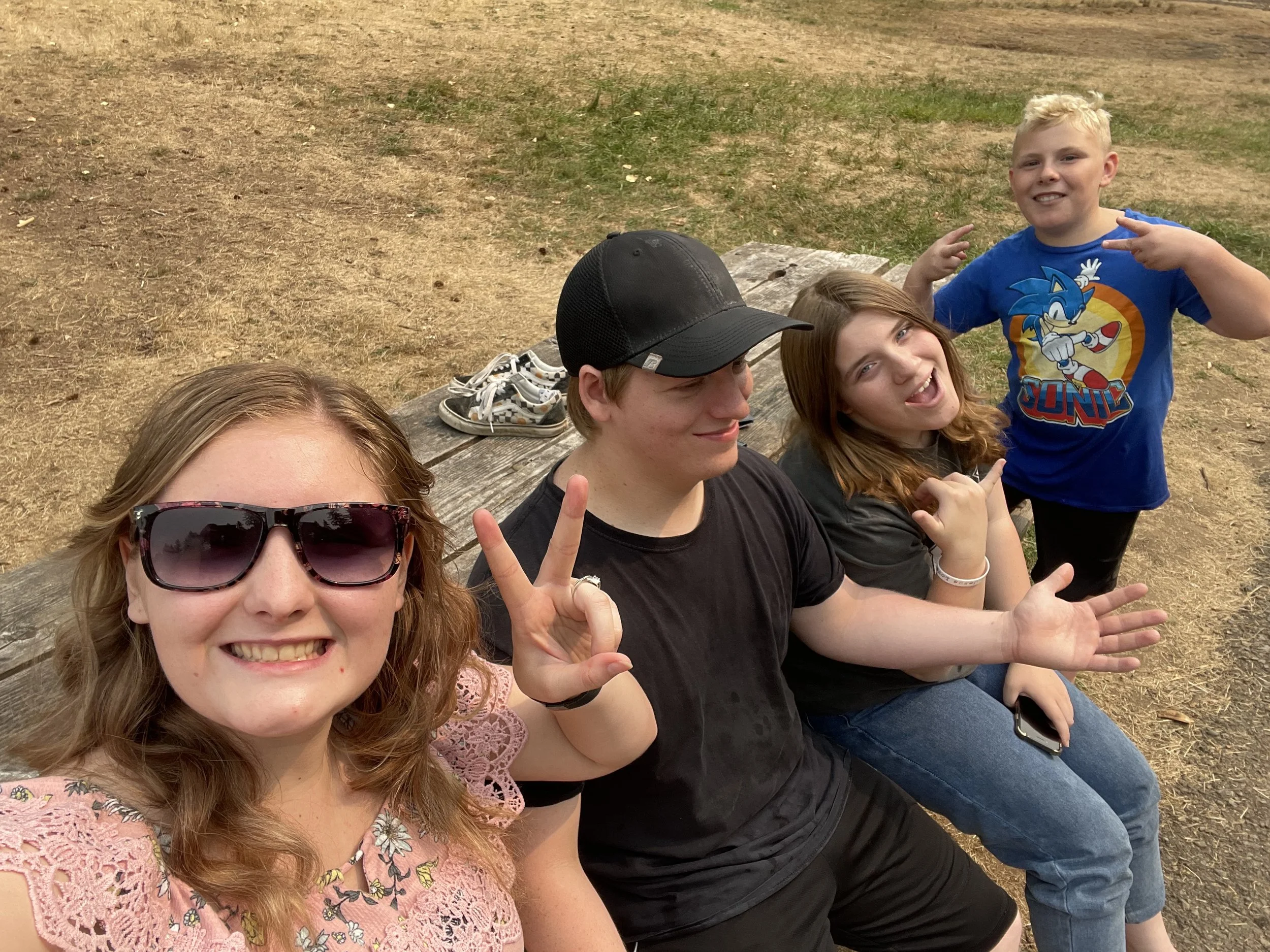 Group of five young friends sitting on a wooden bench outdoors, making playful gestures and smiling, with a dirt and grass background, and shoes on the bench behind them.