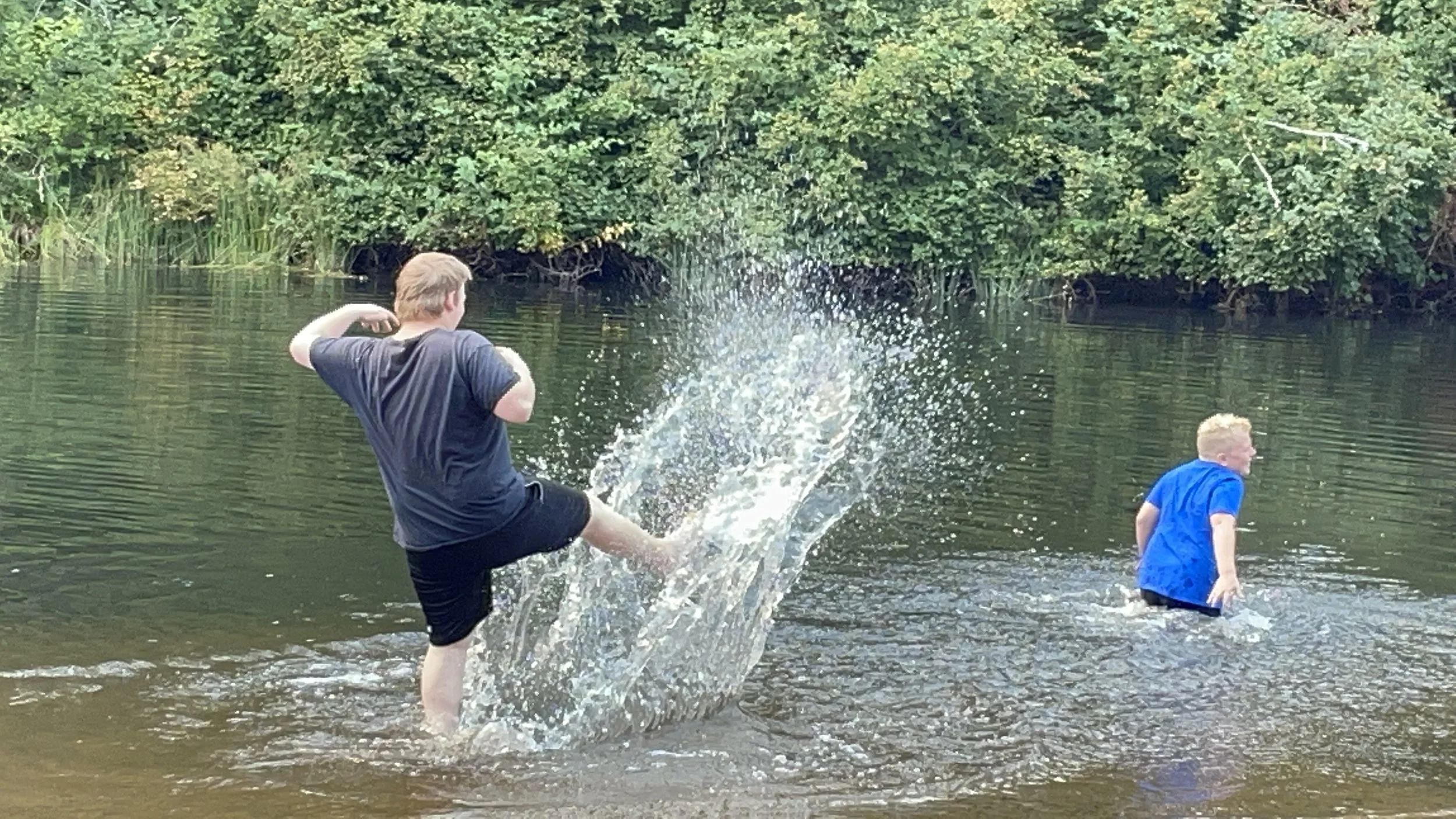 Two boys playing in a shallow river with water splashing as one kicks water towards the other, surrounded by lush green trees.