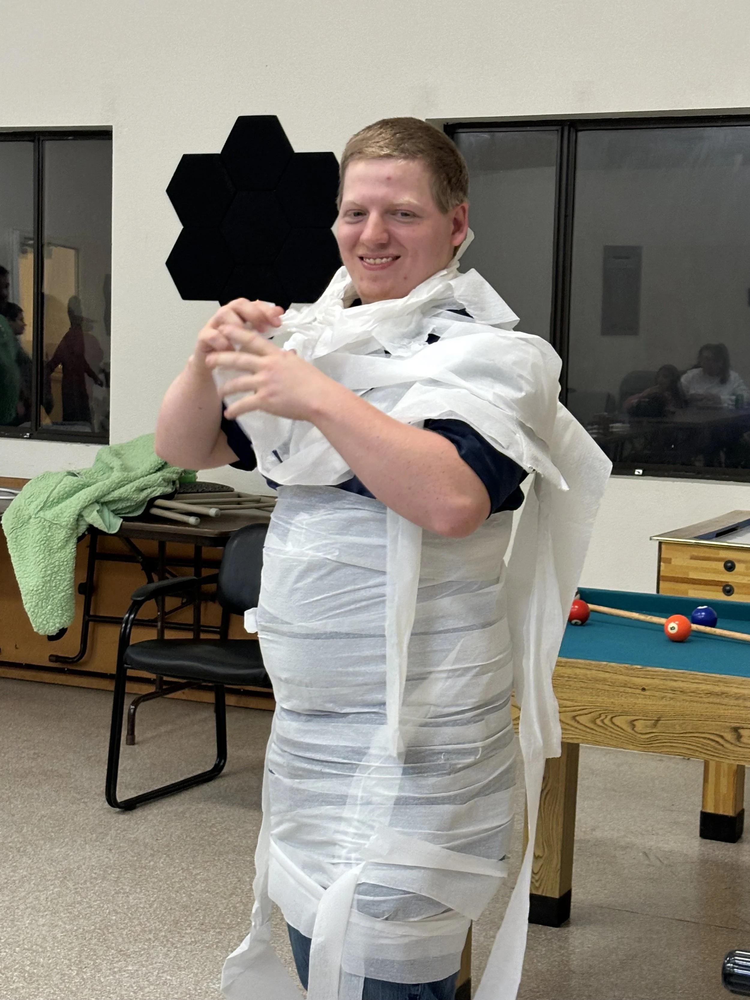 A young man is standing indoors, wrapped in white toilet paper, smiling at the camera.