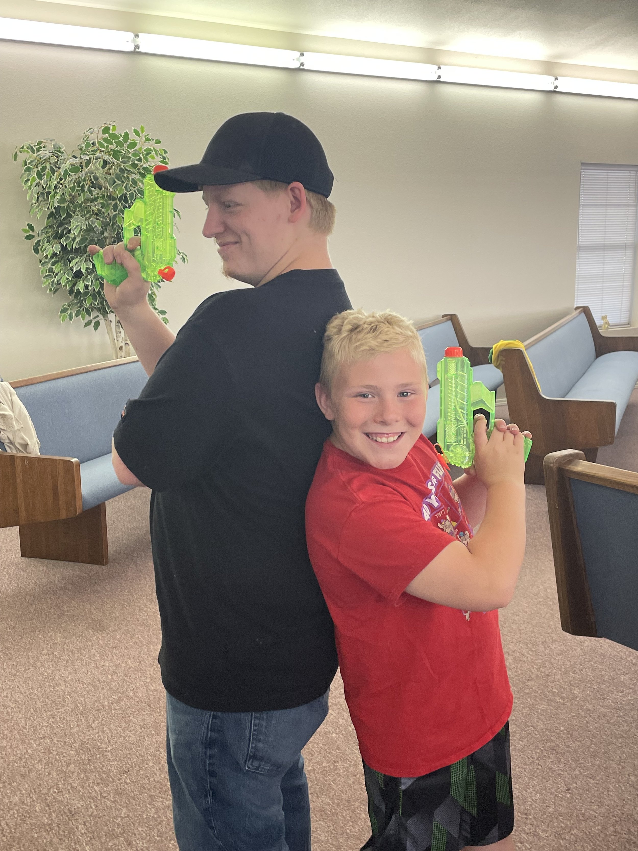 Two boys playing with green water guns back-to-back in a room with beige walls and blue benches.