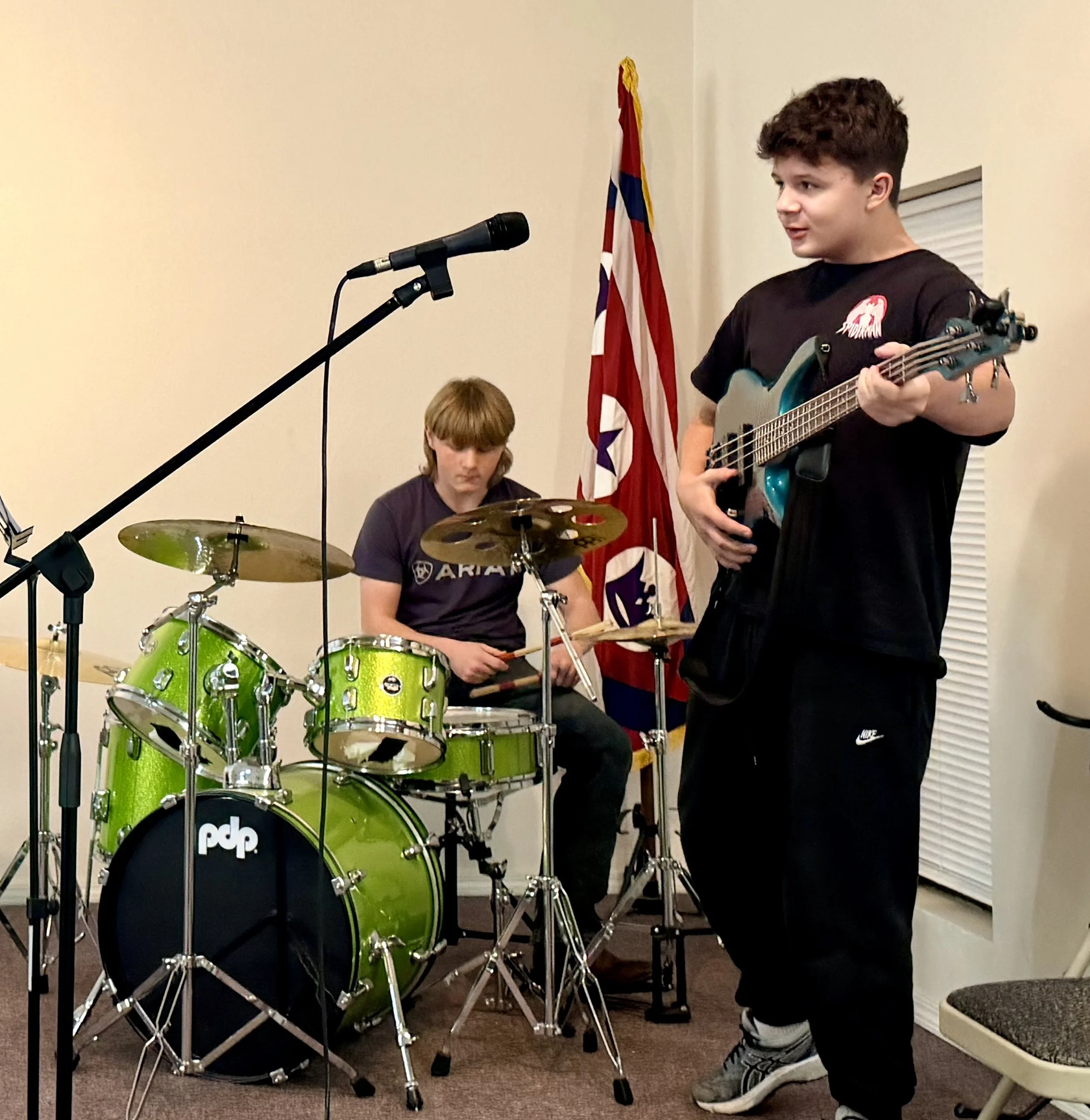 Two boys performing music indoors, one playing drums and the other playing an electric guitar, with a flag in the background.