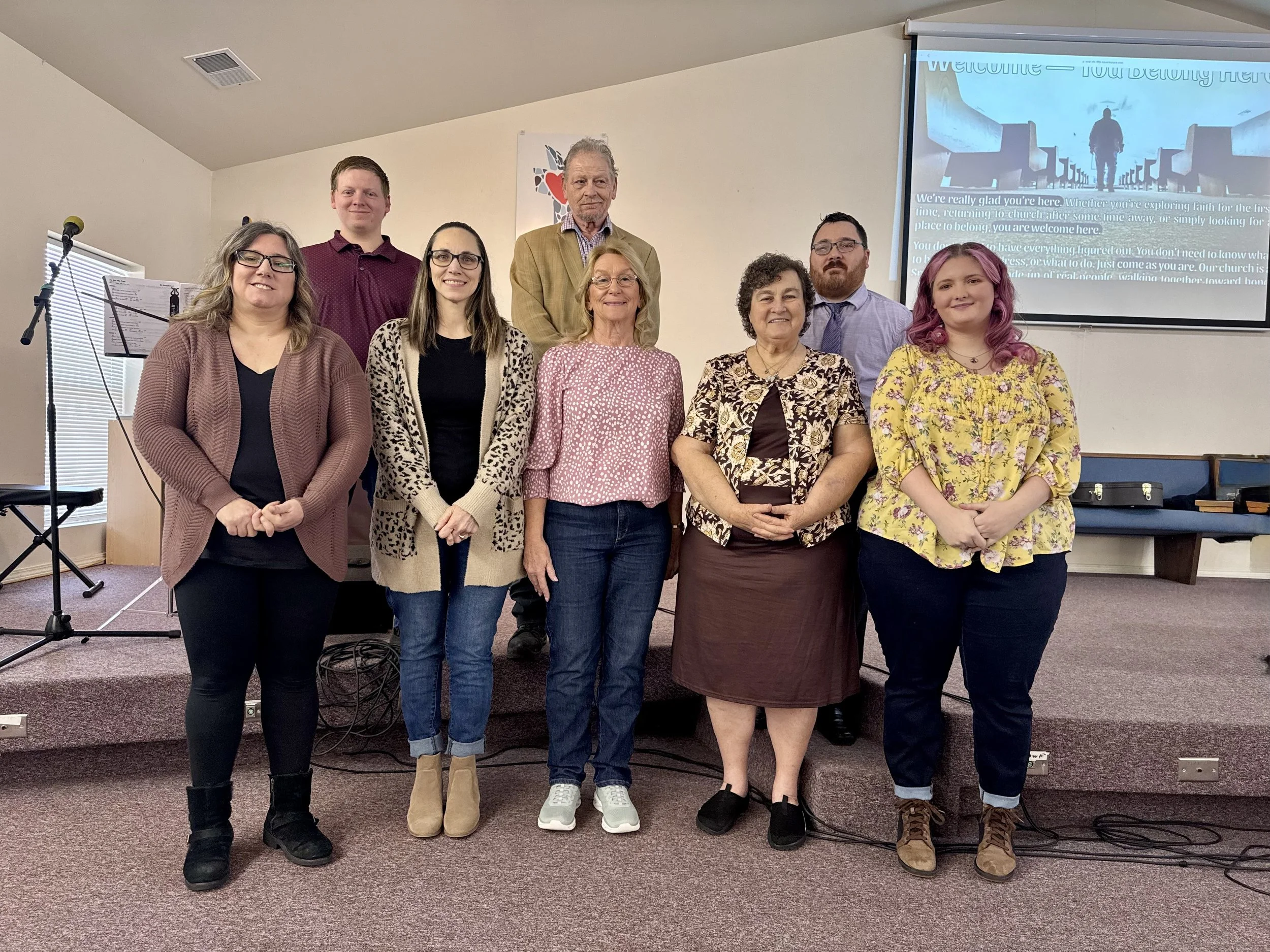 A group of nine people standing in a room with a church presentation slide projected on the wall behind them.