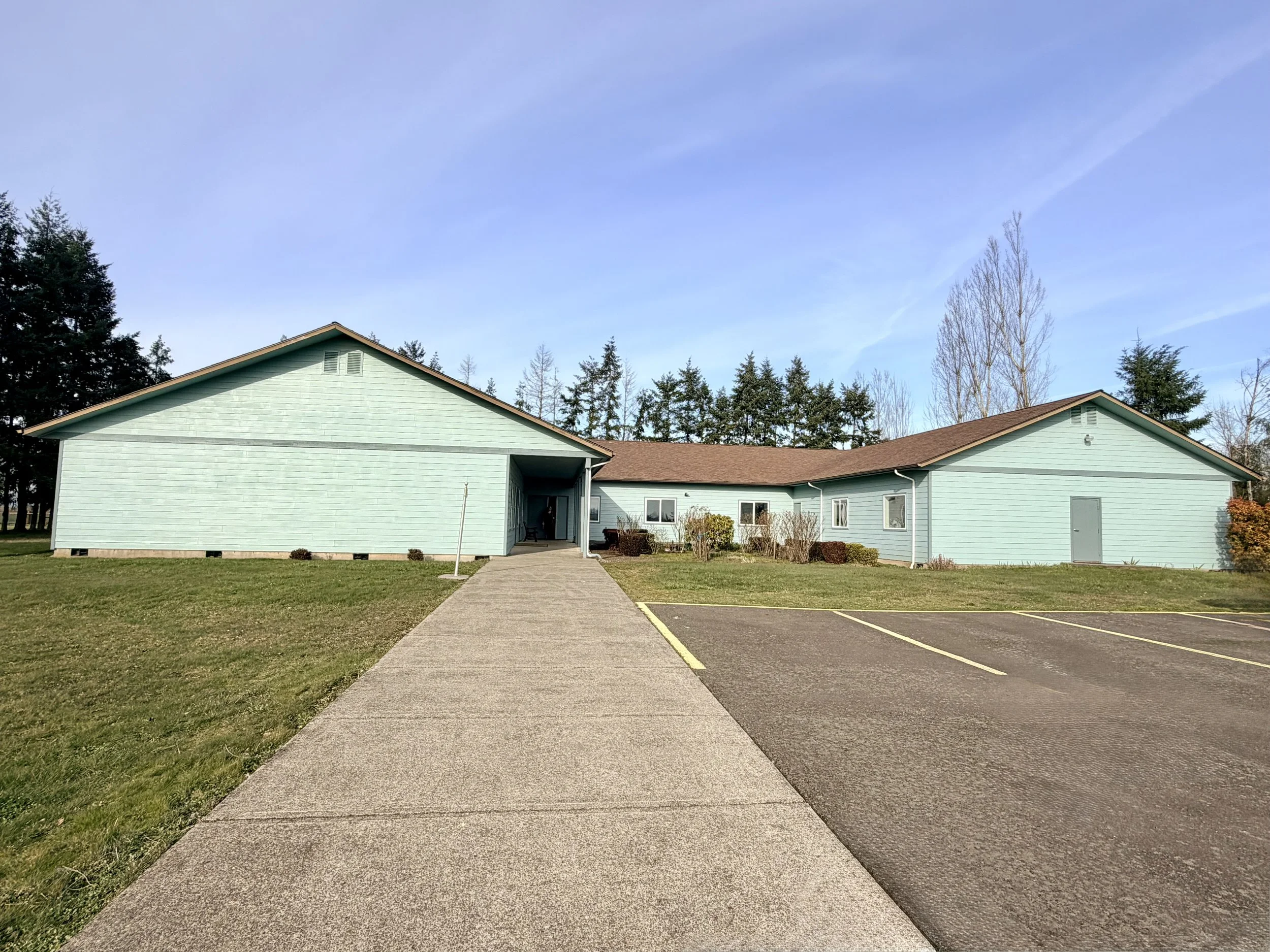 A light blue building with a brown roof, surrounded by grass and shrubs, with a parking lot to the right and a sidewalk leading to the entrance, against a background of trees and a clear sky.