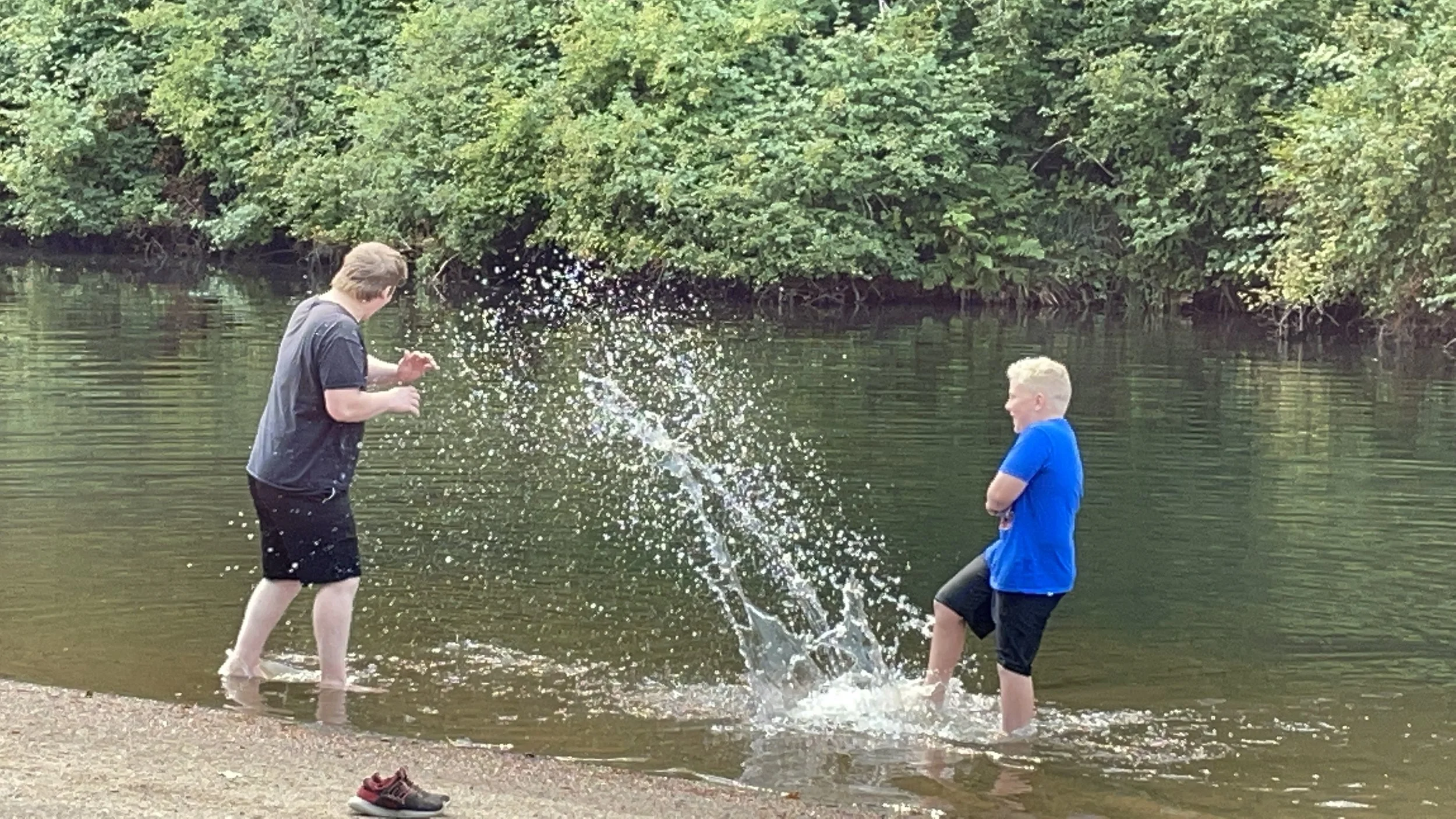 Two boys playing in a shallow river, one splashing water towards the other, with greenery and trees in the background.
