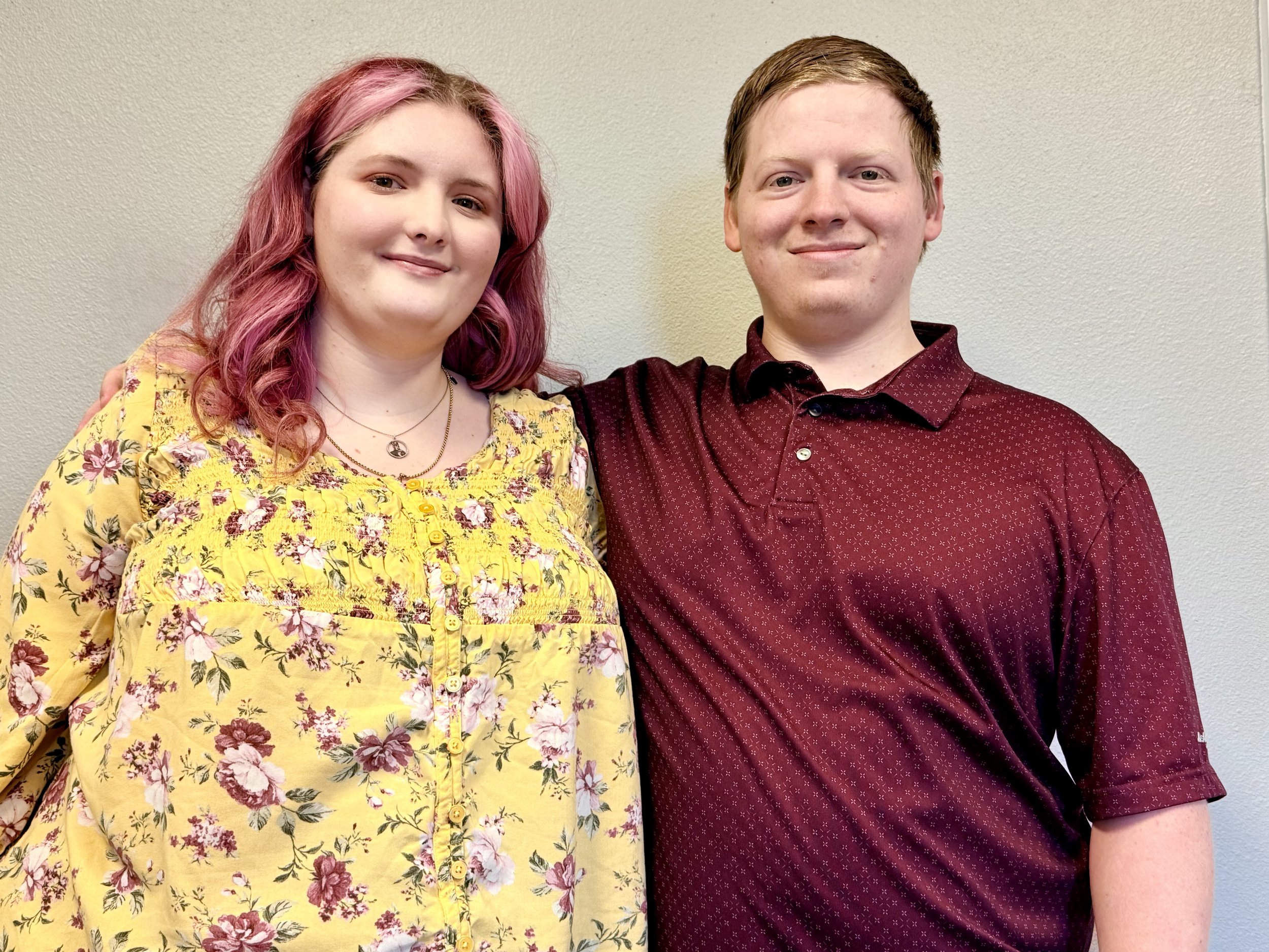 A young woman with pink wavy hair wearing a yellow floral blouse and a young man with short light brown hair in a maroon patterned polo shirt, standing against a plain wall.