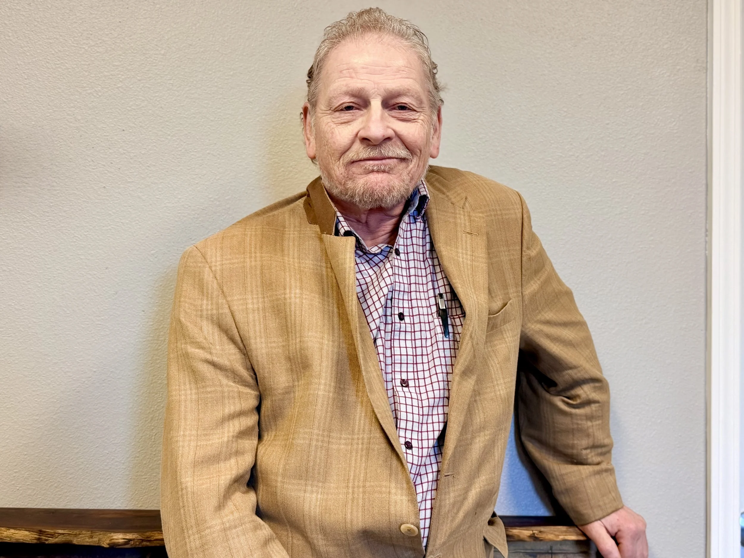 A middle-aged man with light skin, gray hair, and a beard, wearing a tan blazer and a checkered shirt, standing against a beige wall. He is smiling gently and leaning slightly with one hand resting on a wooden surface.