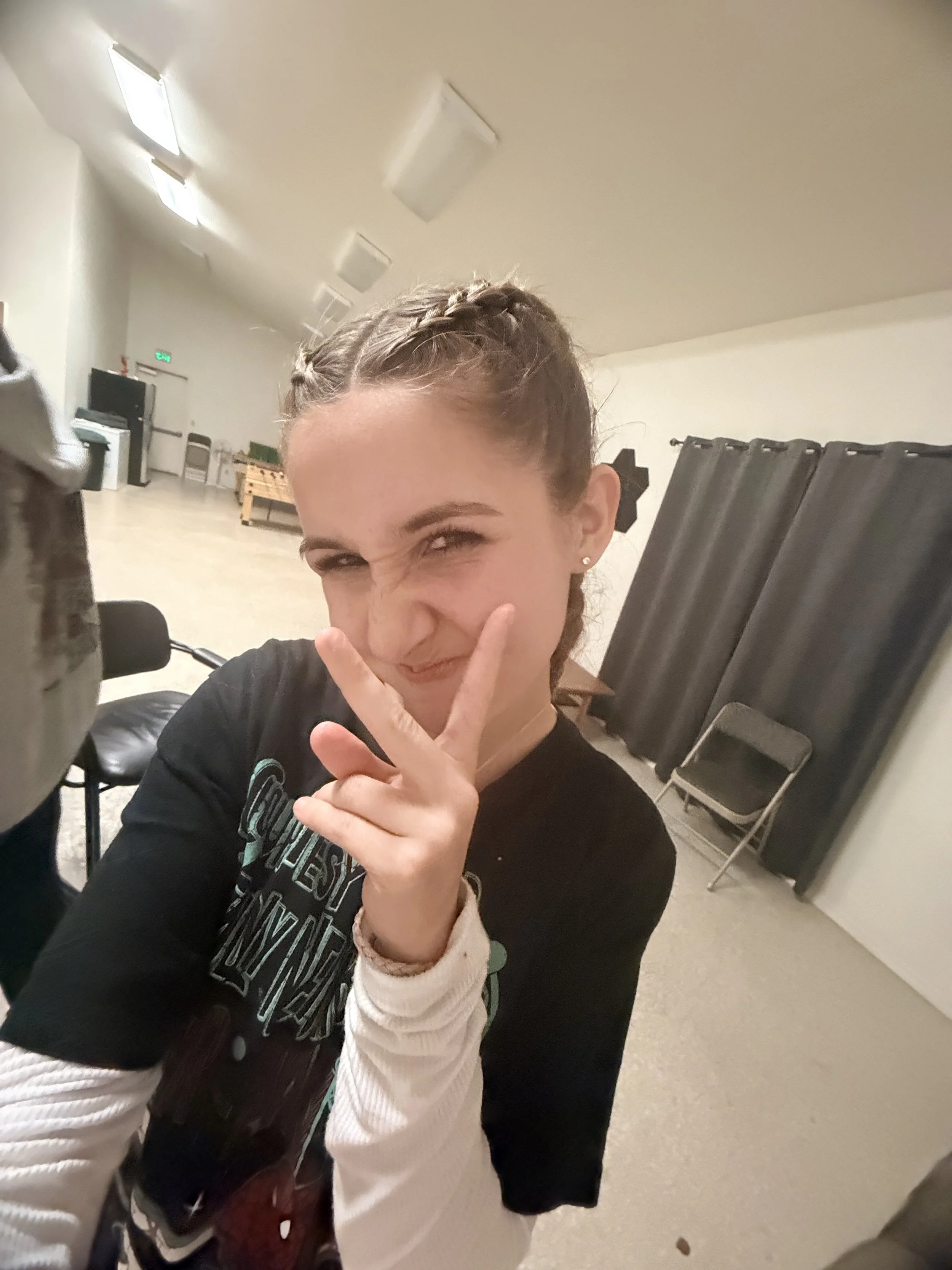 A young woman with braided hair making a peace sign near her face in a large, empty room.