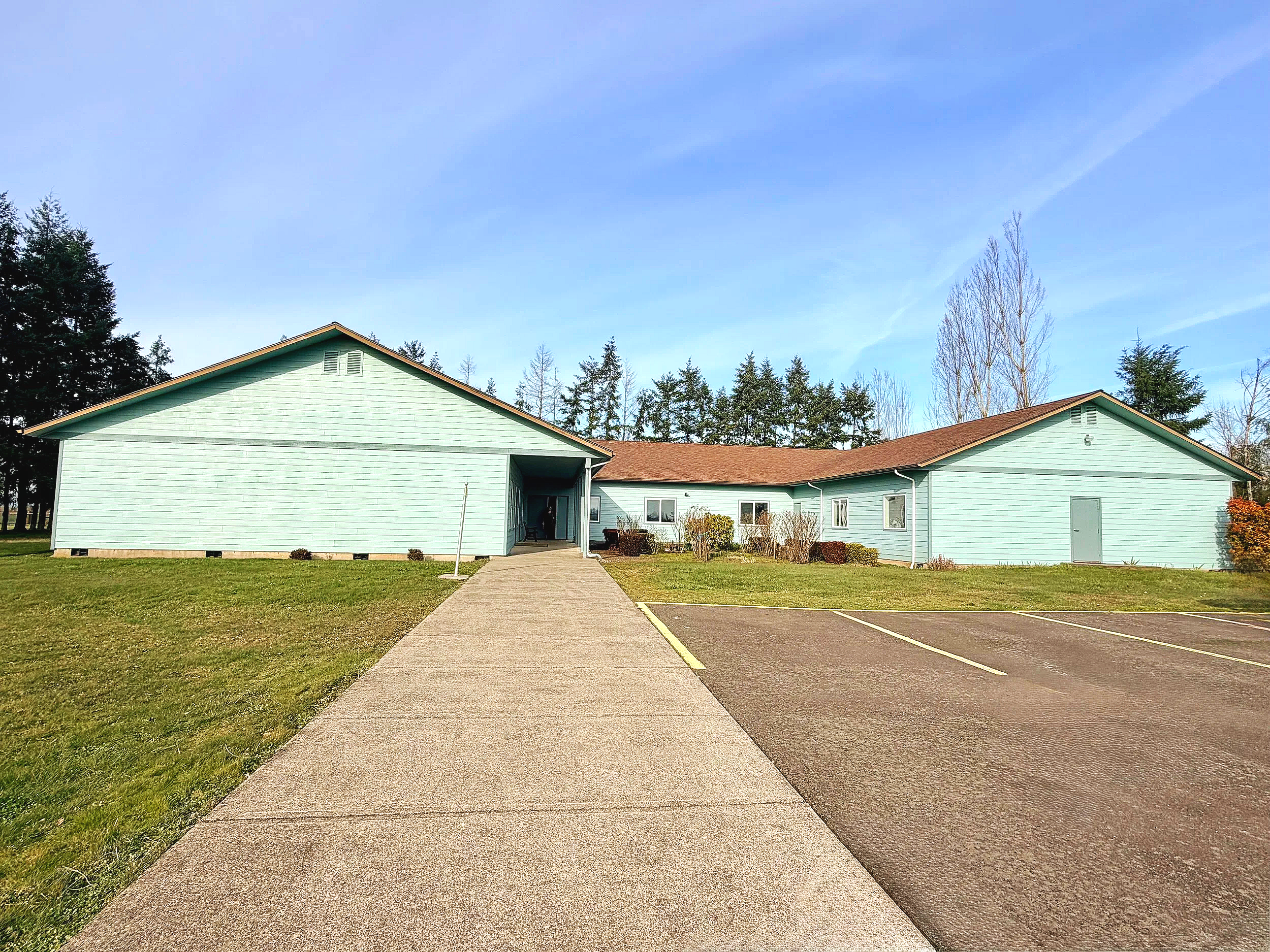 A light blue, single-story building with a pitched roof, surrounded by a grassy lawn, concrete walkway, and parking lot, under a clear blue sky.