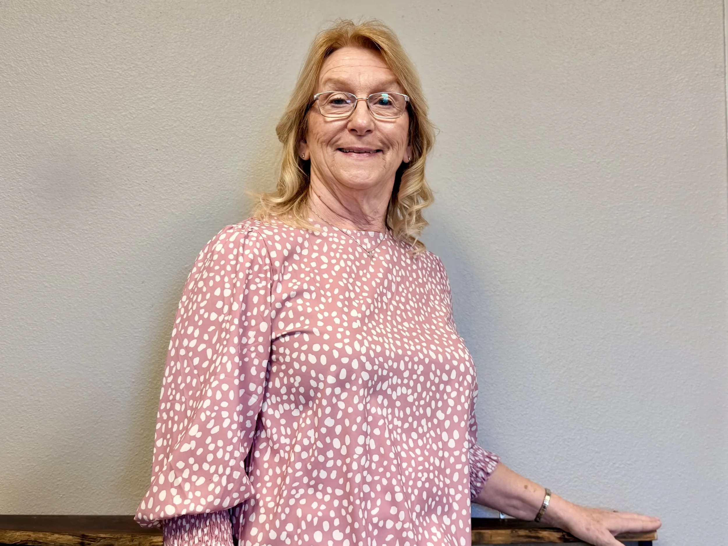 A smiling middle-aged woman with glasses and blonde hair, wearing a pink dress with white irregular dot pattern, standing against a beige wall.