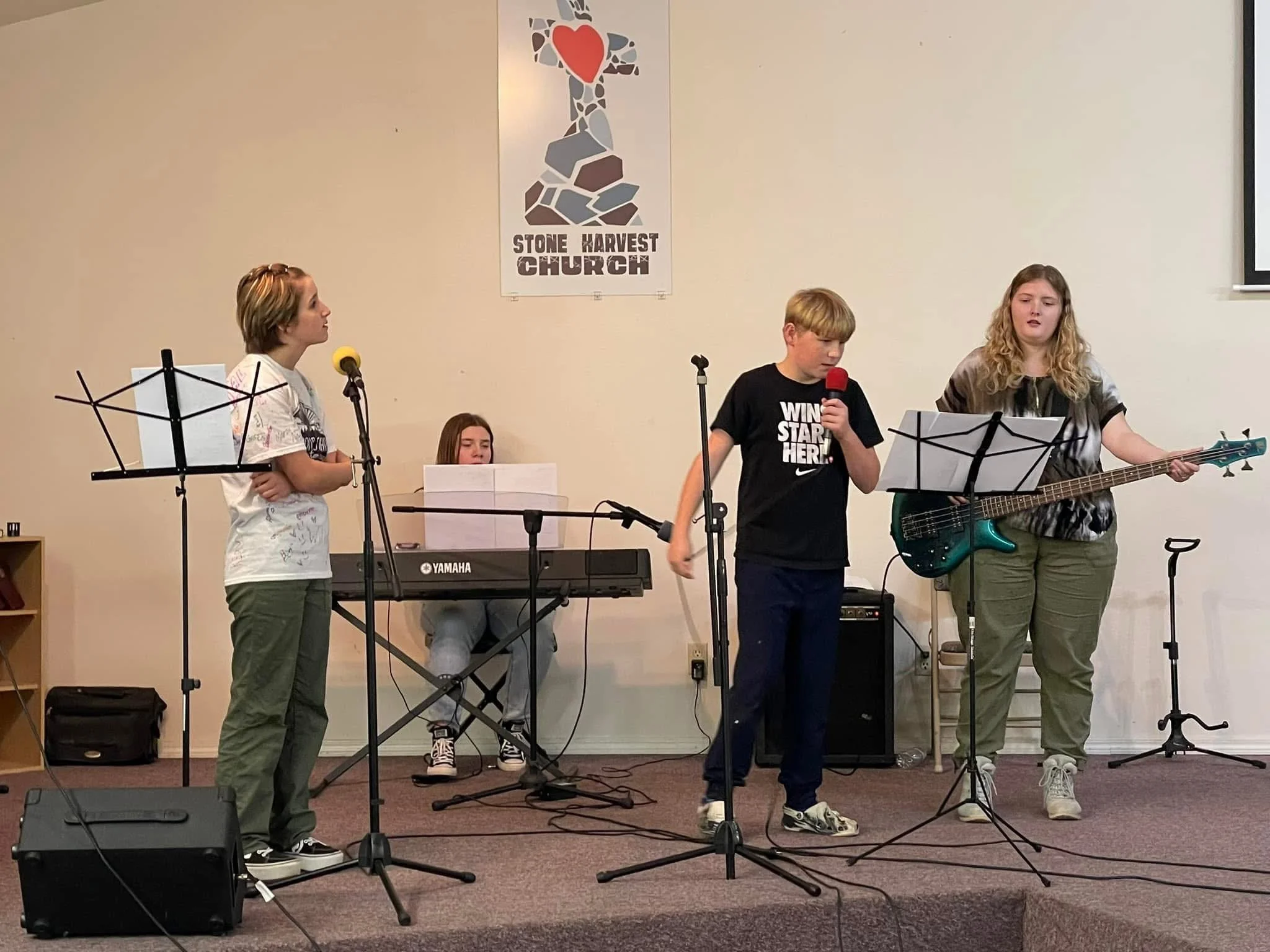 A group of four children performing music on a stage at Stone Harvest Church with musical instruments including a keyboard, microphone, and bass guitar.
