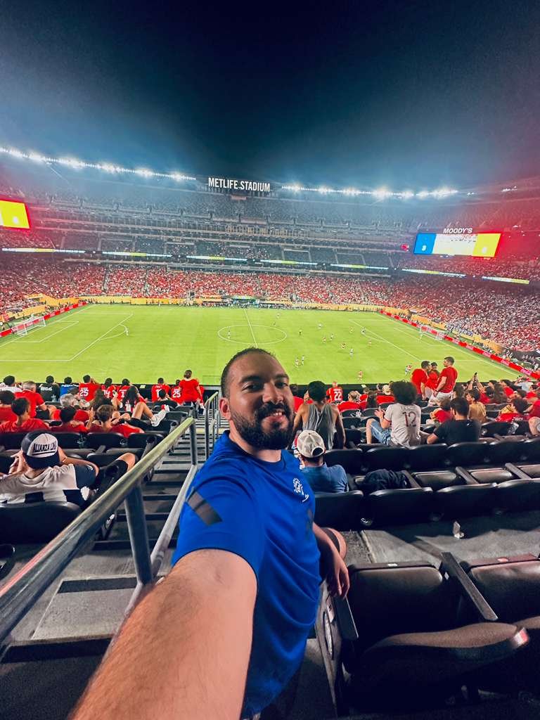 Nour Mohsen taking a selfie at a soccer stadium filled with spectators watching a game on the field, with the stadium lights on at night.