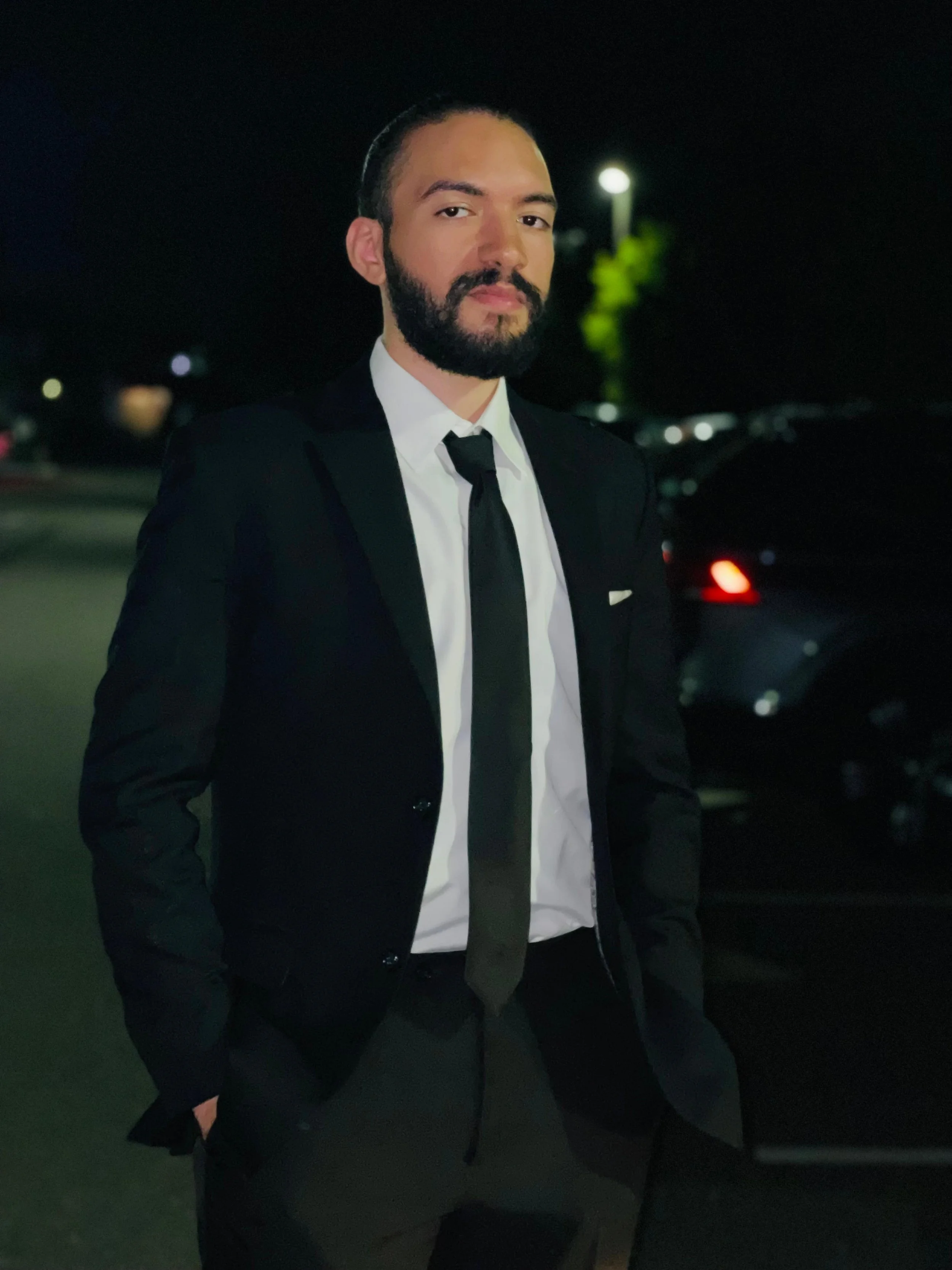 Nour Mohsen in a black suit, white shirt, and black tie standing outdoors at night, with cars and streetlights in the background.