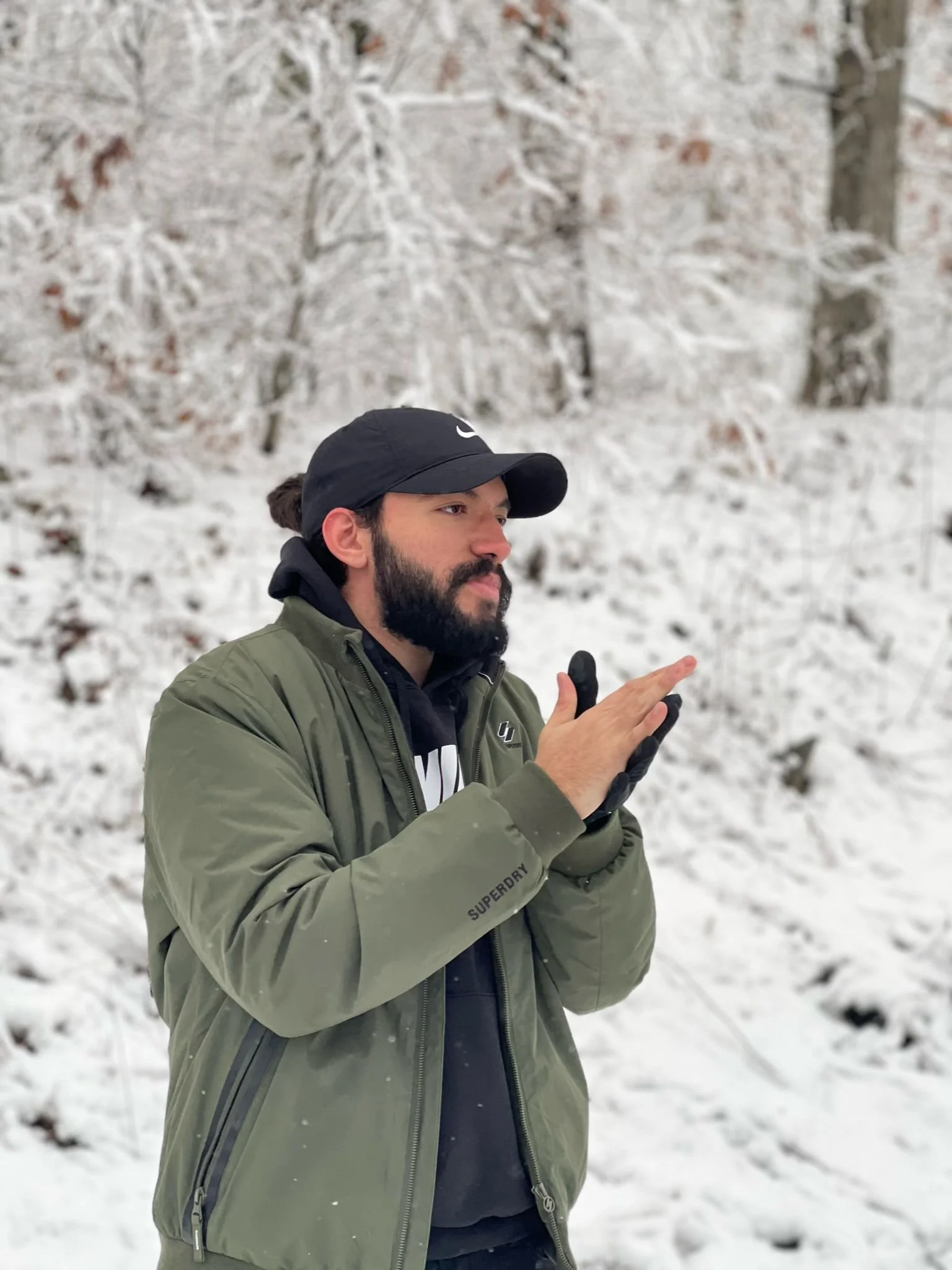 Nour Mohsen with a beard, wearing a black cap, green jacket, and gloves, standing in a snowy forest, looking at his hand.
