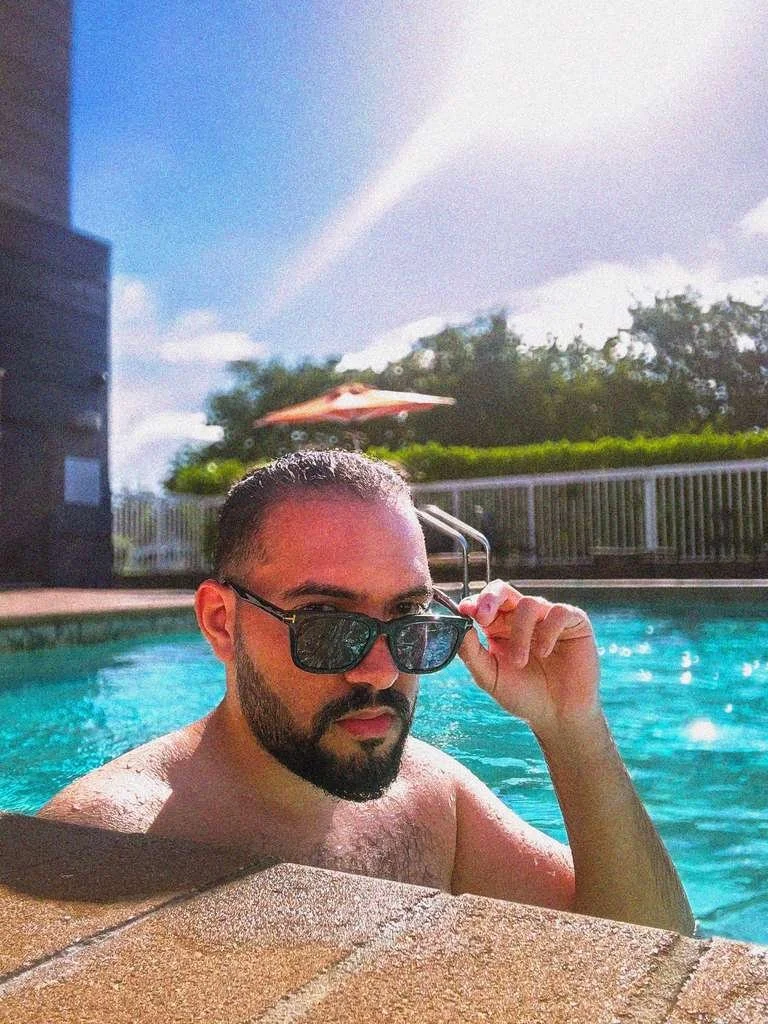 Nour Mohsen with a beard and sunglasses sitting in a swimming pool adjusting his sunglasses with a fence, greenery, an umbrella, and a blue sky with a rainbow in the background.
