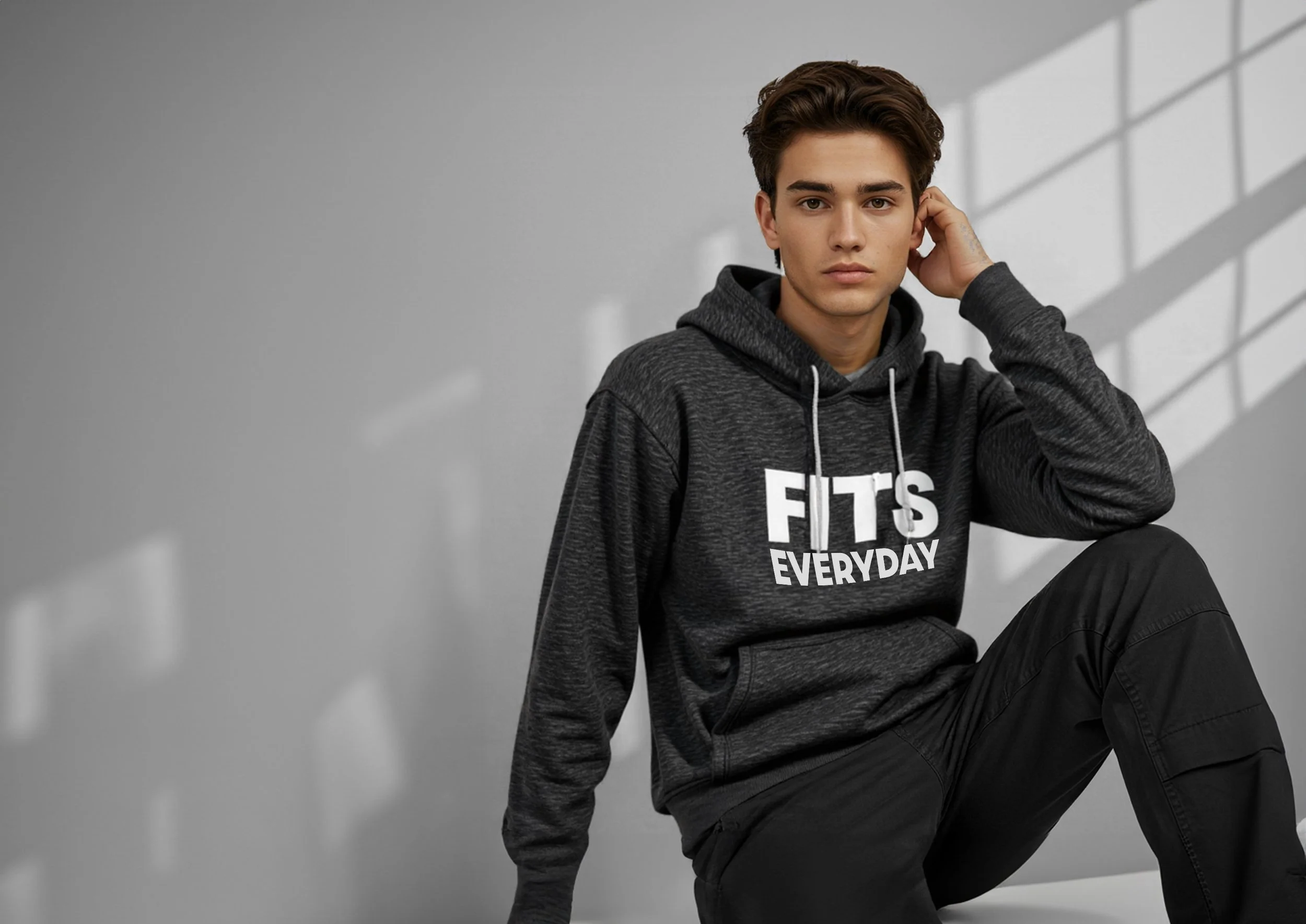 A young man with dark hair wearing a black hoodie with the text 'FITS EVERYDAY' sitting against a gray wall with shadows from a window.