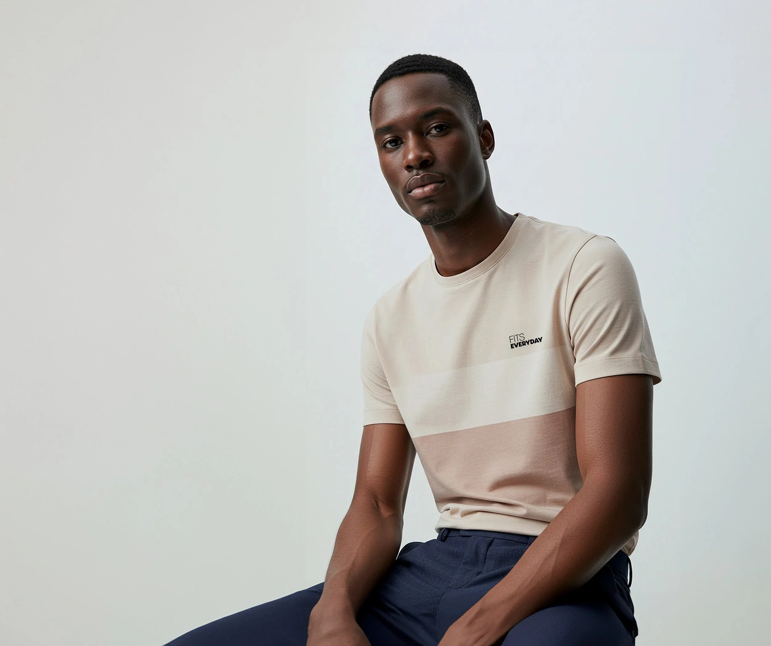 A man sitting against a plain light background, wearing a beige t-shirt with the words 'FITS EVERYDAY' on the chest, and navy pants.