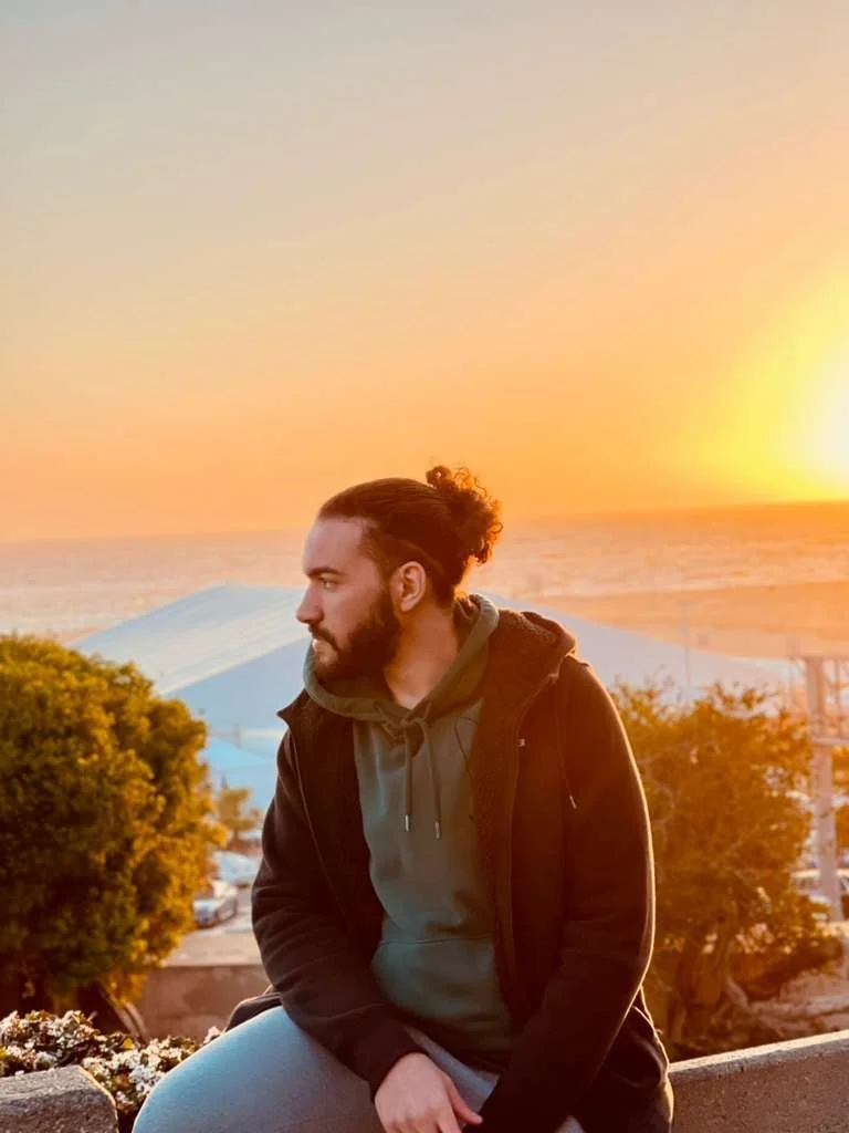 Nour Mohsen with a beard and long hair tied back, wearing a hoodie and jacket, sits outdoors during sunset by the ocean, with trees and umbrellas in the background.