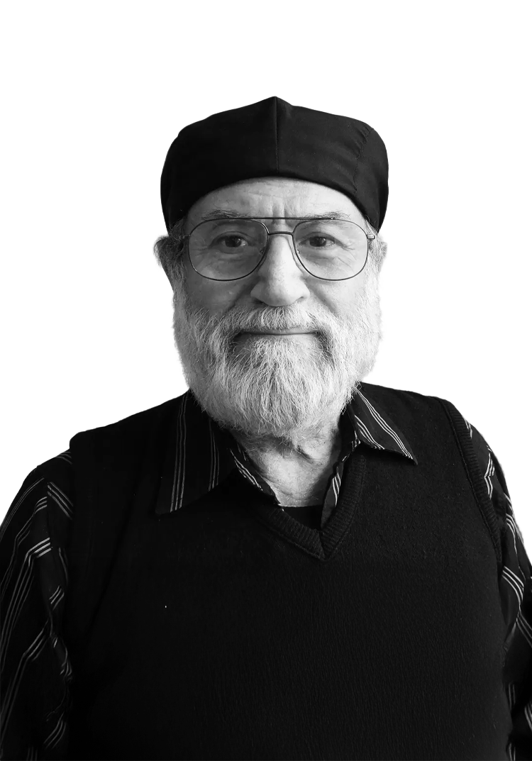 An elderly man with glasses, a beard, and a hat, wearing a sweater over a collared shirt, smiling at the camera.