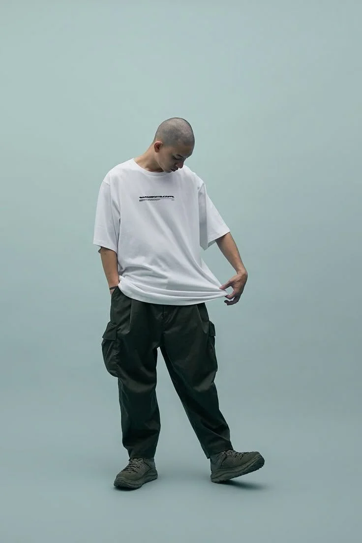 Person standing against a plain light background, wearing a white oversized T-shirt, black cargo pants, and brown shoes, looking down and touching the hem of the shirt.
