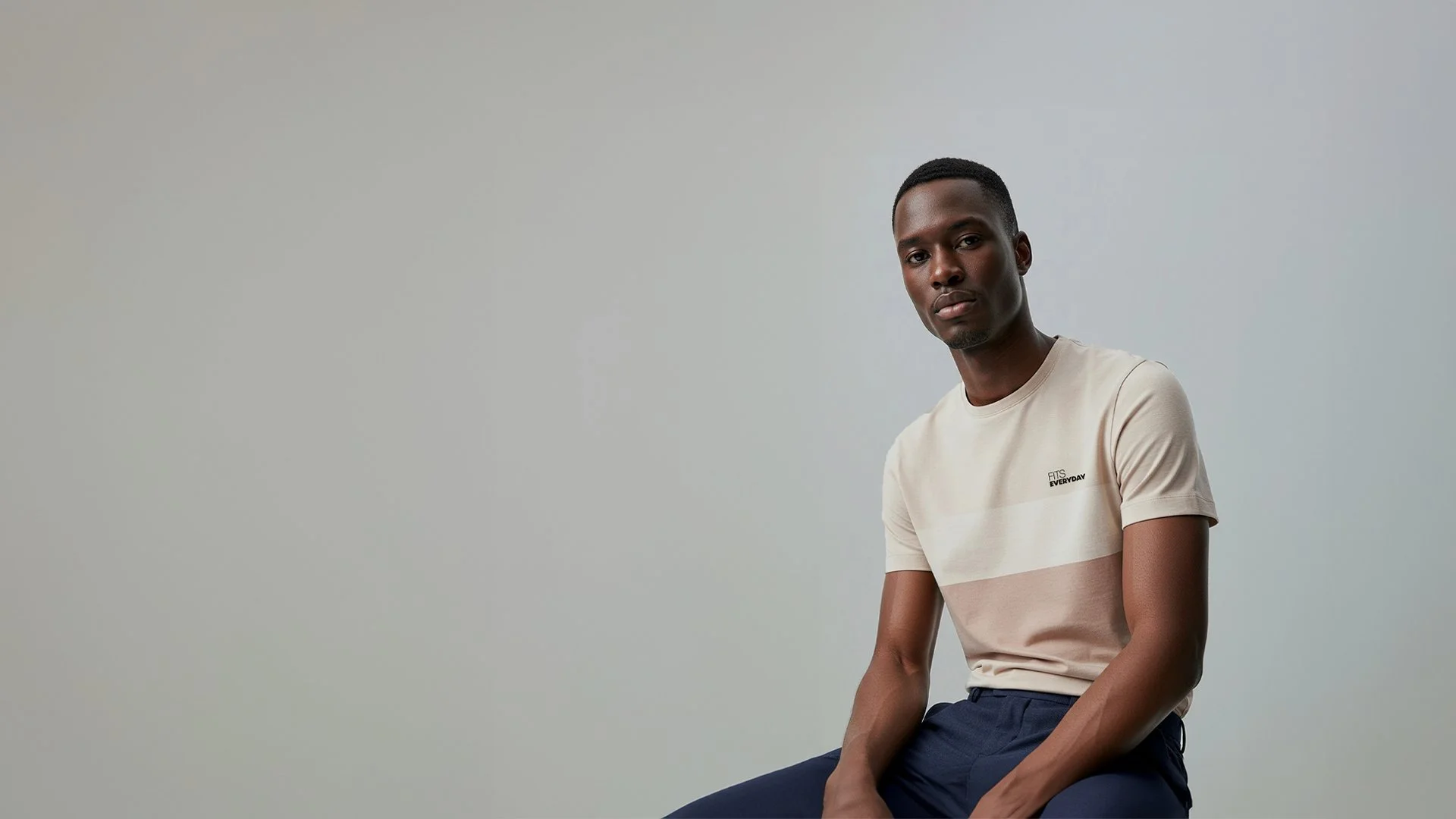 A young man with dark skin and short hair sitting against a plain light gray background, looking directly at the camera with a neutral expression.
