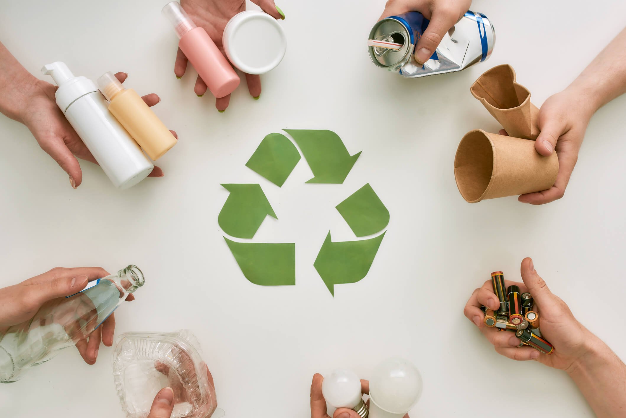 Hands holding various recyclable items such as plastic bottles, aluminum cans, glass bottles, paper cups, biodegradable cups, and batteries arranged around a green recycling symbol on a white surface.
