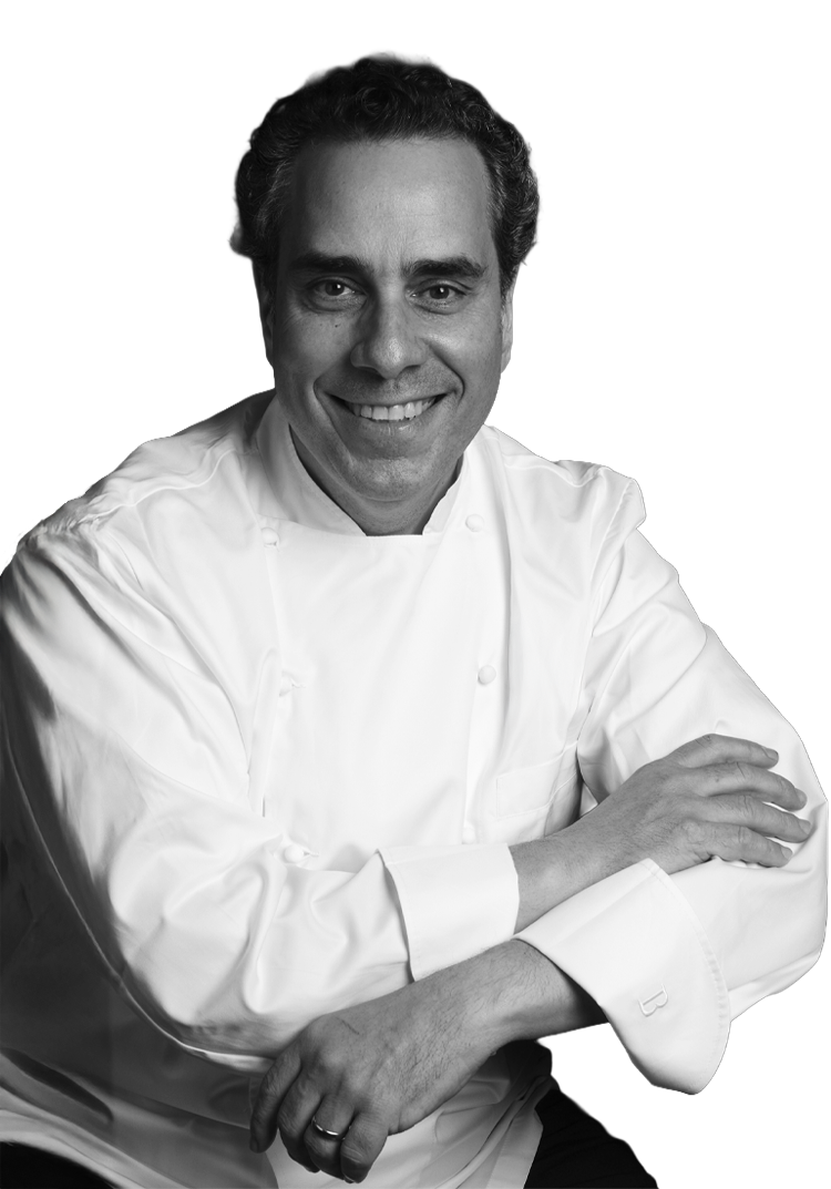 Black and white photo of a smiling male chef in a white chef's coat, sitting with his arms crossed.