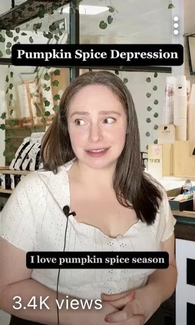 A woman with long brown hair and a white top, sitting in front of a shop display, looking to the side with a slight smile. The background includes shelves and decor, with text overlay saying 'Pumpkin Spice Depression'.